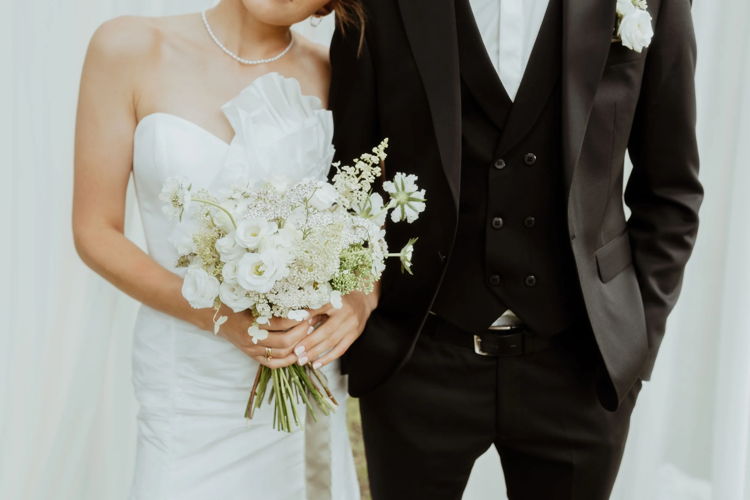 Bride in a white wedding dress holding a bouquet of white flowers, standing next to groom in a black tuxedo with a boutonniere.