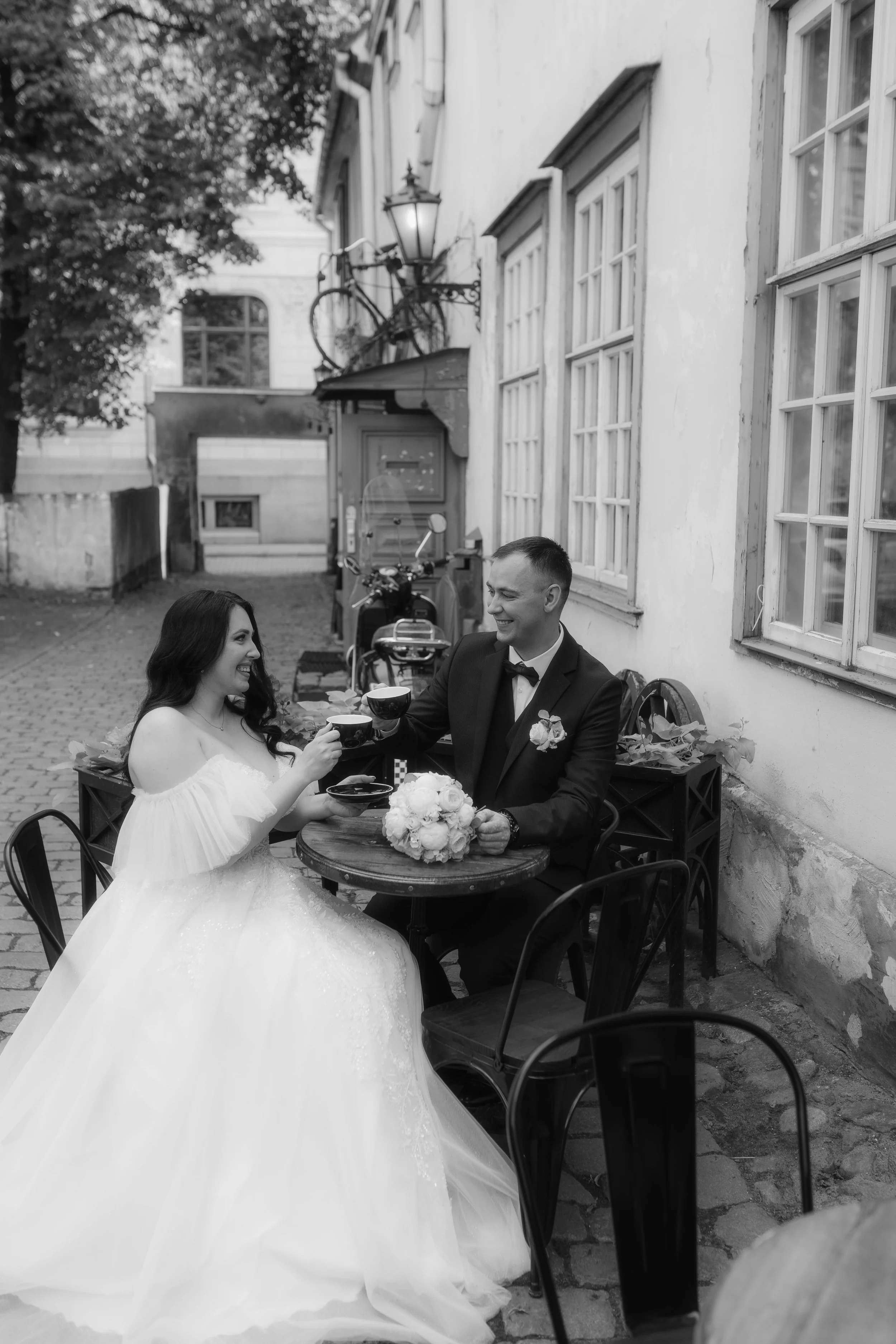 A bride and groom in wedding attire sharing a toast at an outdoor cafe, with the bride wearing a white wedding dress and the groom in a black tuxedo, sitting at a small round table with a bouquet of white flowers.