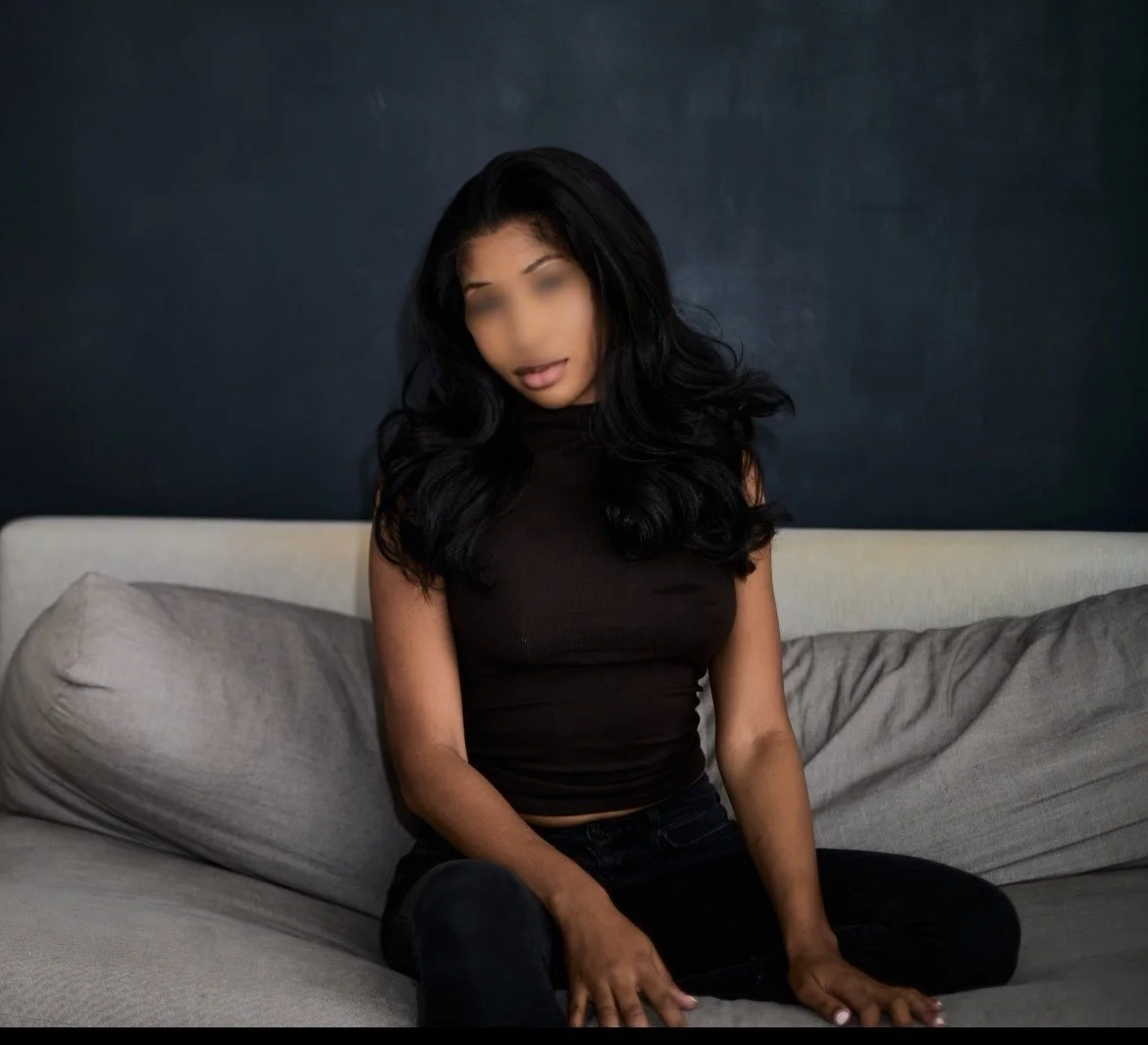 A woman with long black hair sitting on a beige couch with gray pillows against a dark wall, wearing a black top.