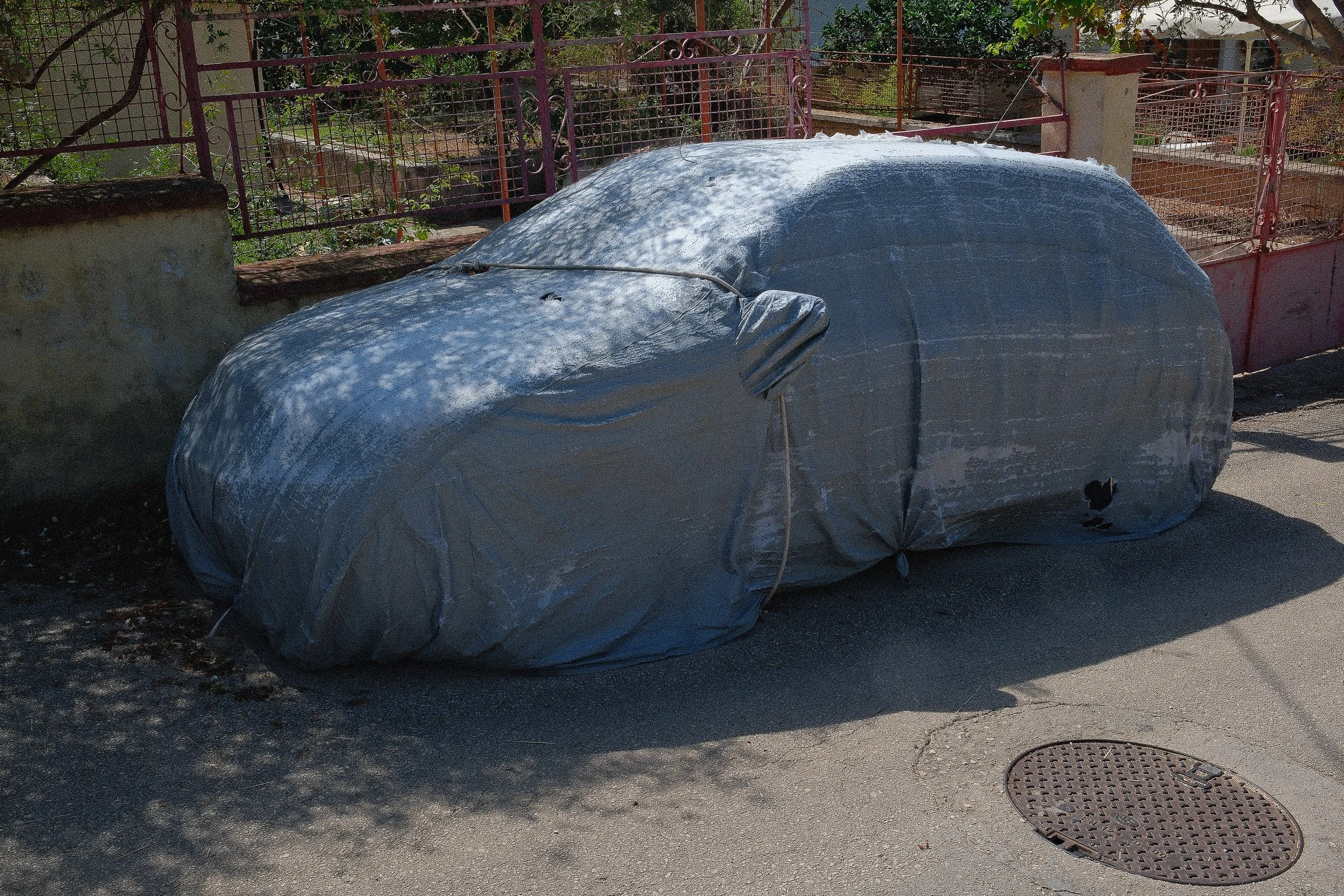 A car covered with a grey protective cover parked on the side of a street near a fence with trees and plants in the background.