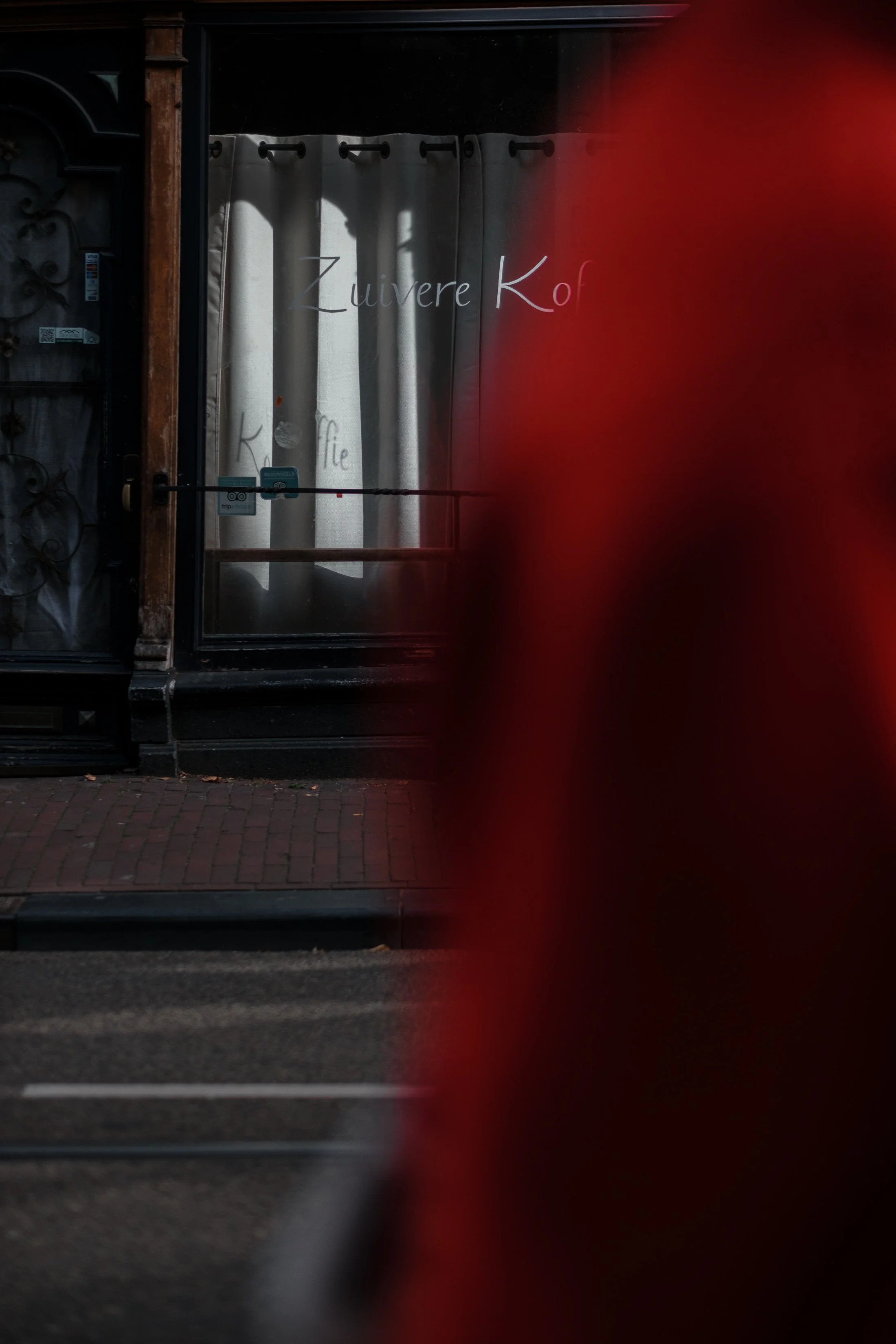 View of a storefront with a curtain, with words "Zuverre Kort" and "Koffie" written on the window, partially obscured by a blurry red object in the foreground.