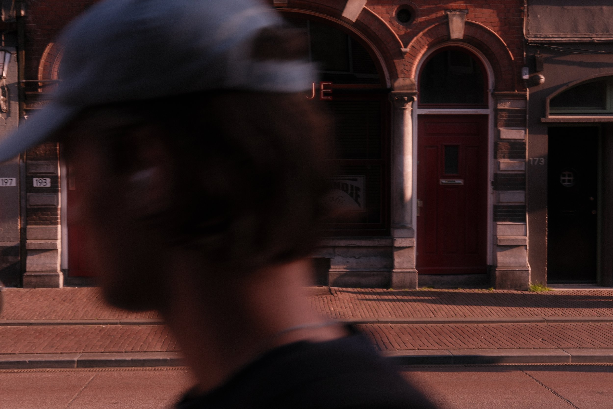 Blurred side profile of a person wearing a cap, walking in front of a brick building with arched windows and red door.