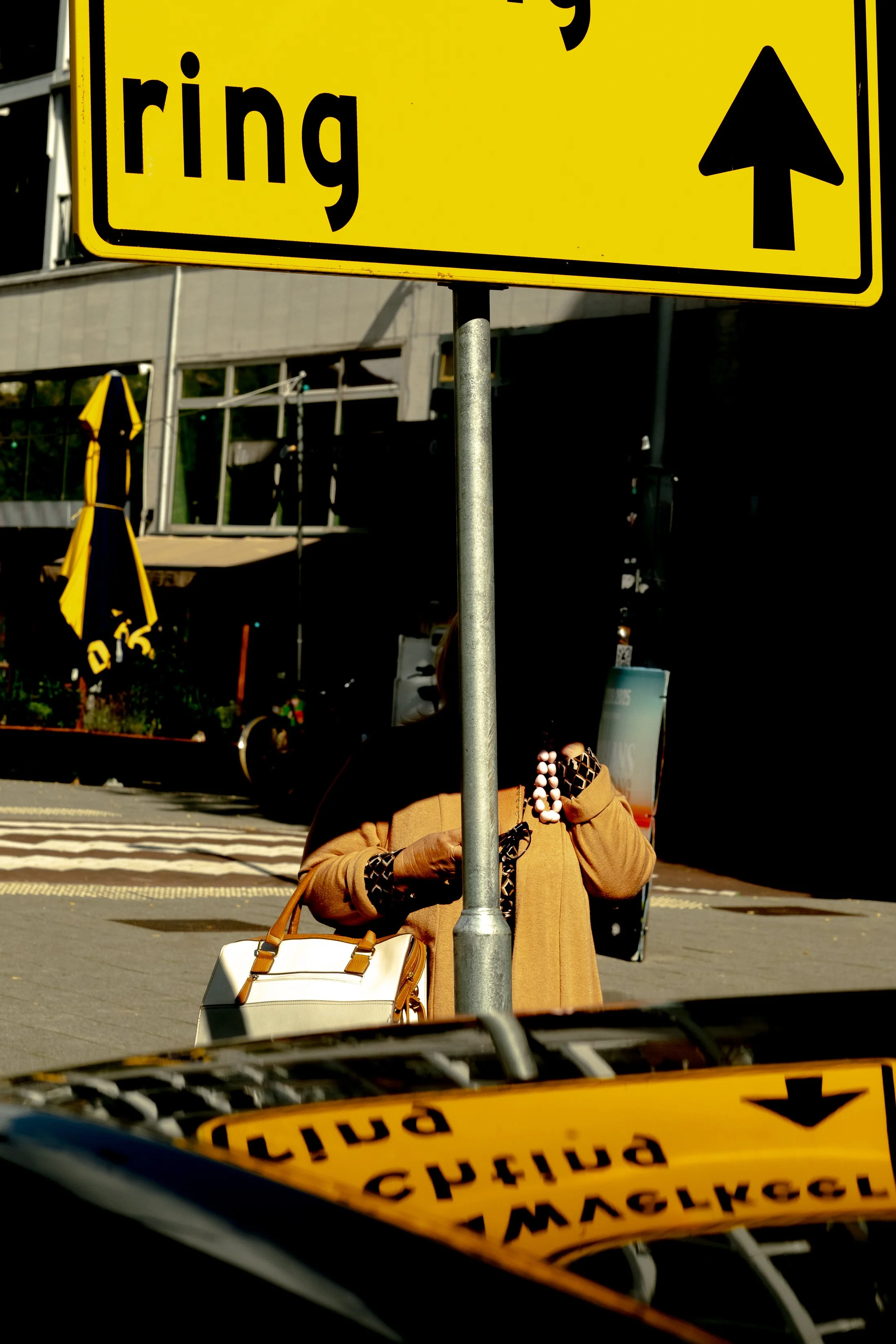 City street scene with a yellow street sign in the foreground, a person in a tan coat and pearl necklace partially obscured behind a pole, carrying a white handbag, and a crosswalk in the background.