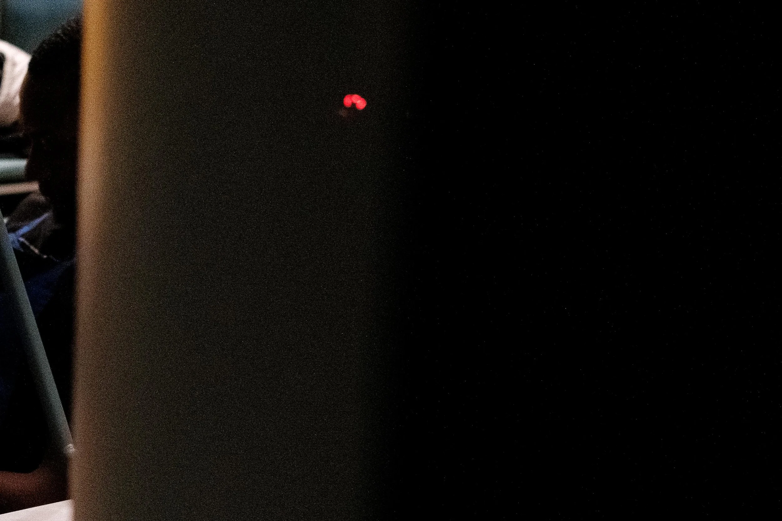 Dark image with a small, blurred red light in the background, a laptop partly covered by a person on the left side, and a blurred background with some objects on the left.