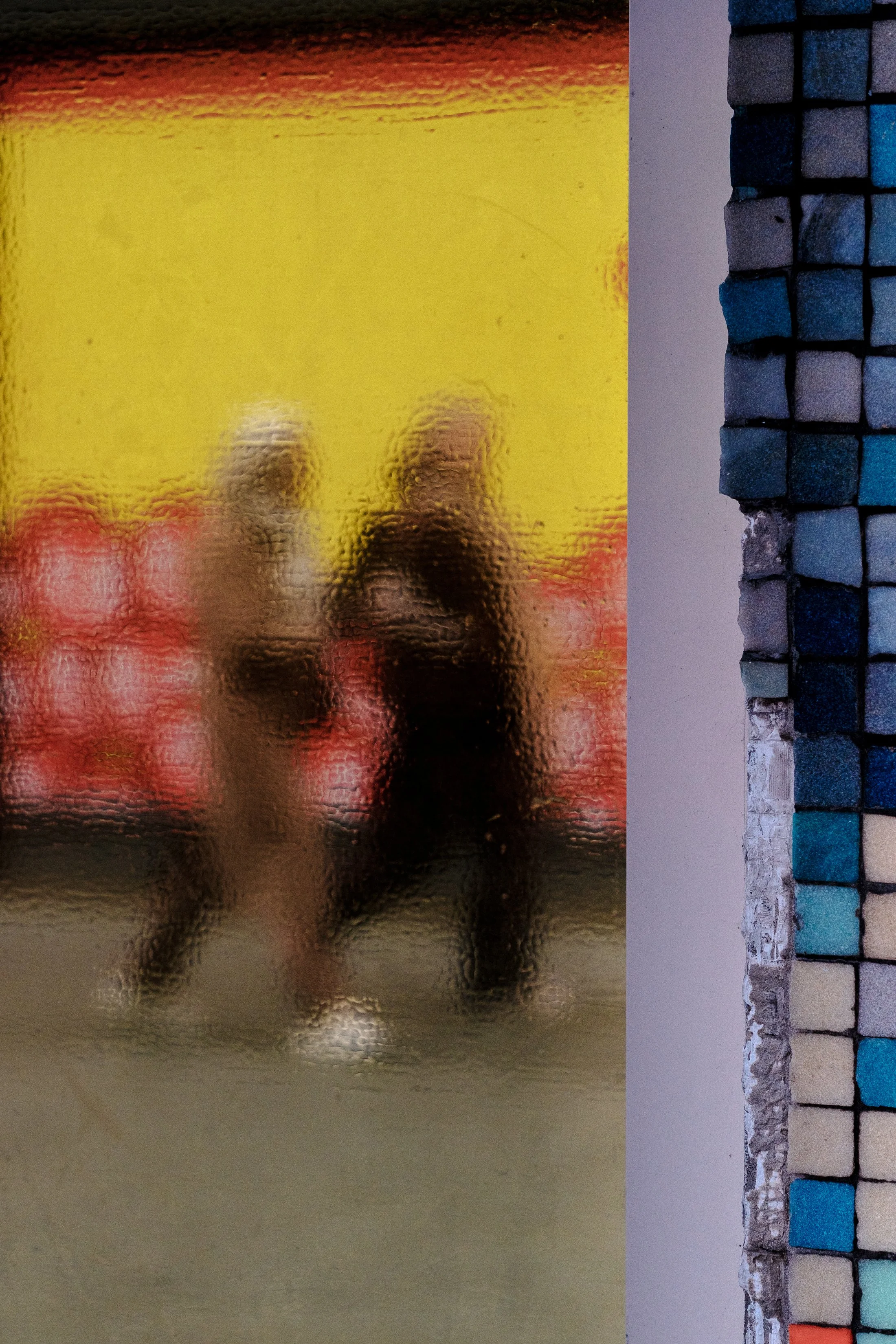 Blurred reflection of a person taking a photo, seen through textured glass, with colorful tiles and a wall on the right side.
