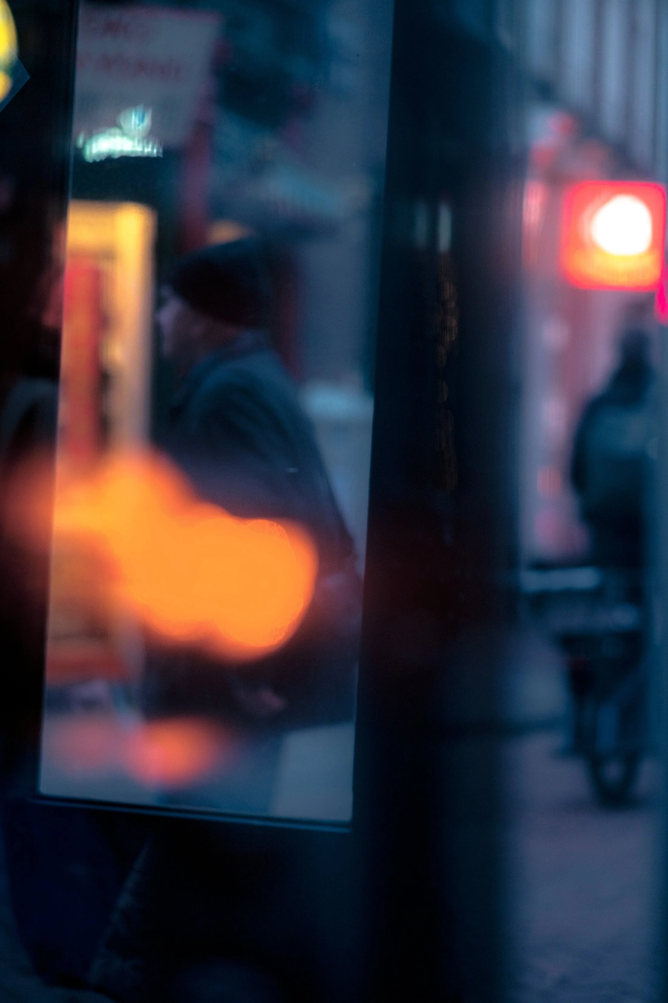 Blurred reflection of person with a mask on a glass window, outdoor scene with neon lights and a person riding a bicycle at night.