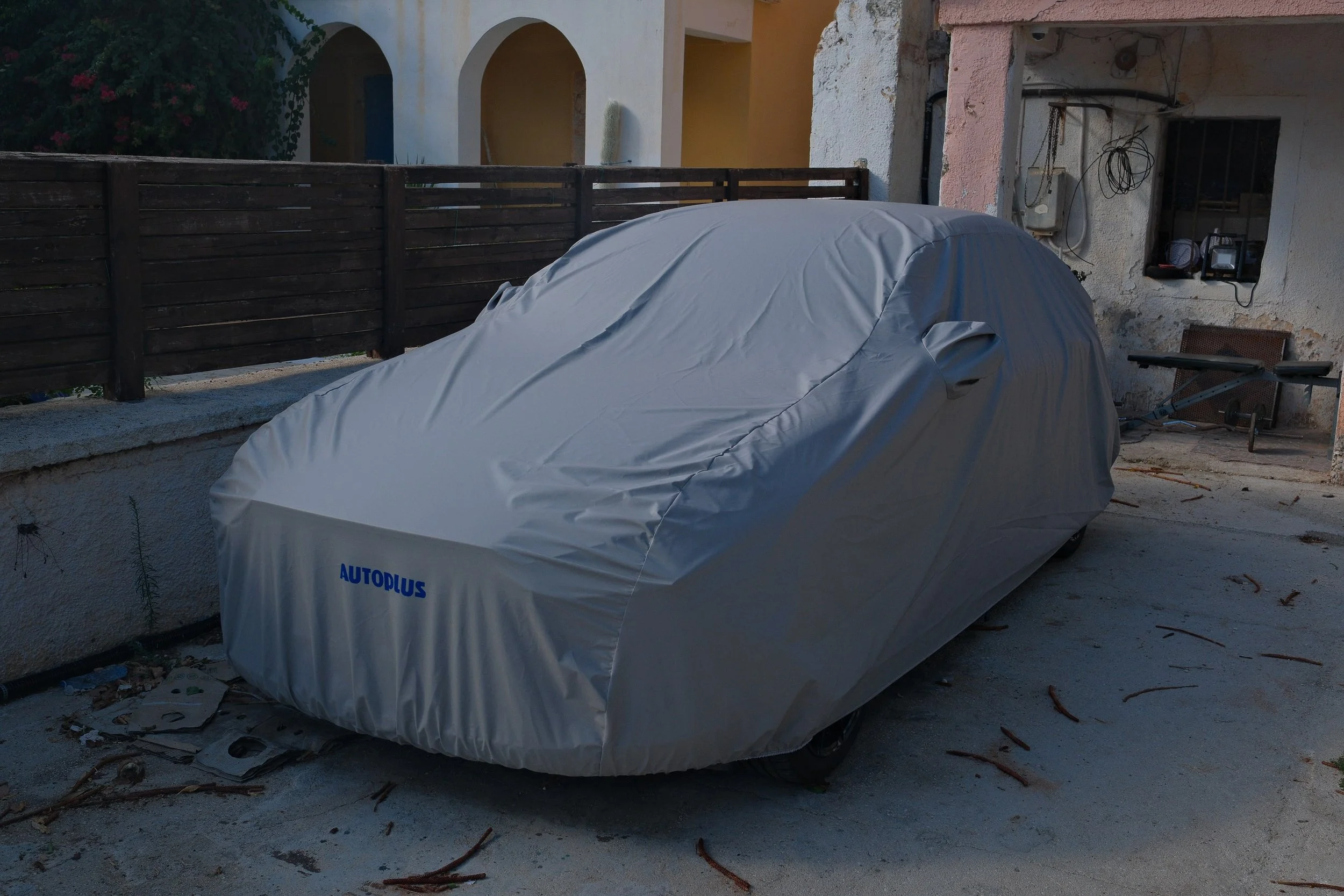 Car covered with a grey car cover labeled 'AUTOPLUS' parked outdoors on a concrete surface next to a low wall and a wooden fence, with a building and a bush with pink flowers in the background.