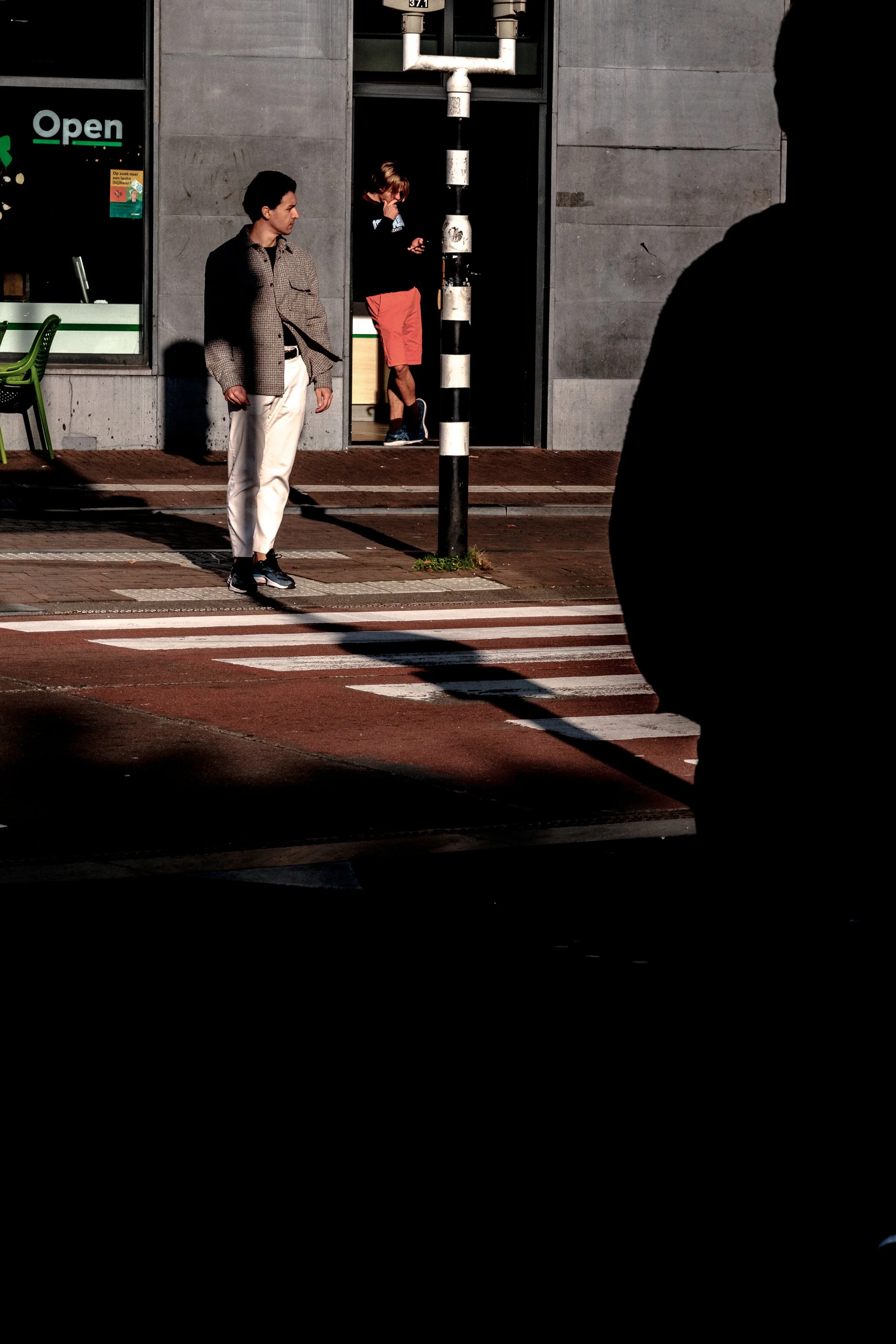 Two men waiting at a crosswalk, with one man on the street and another visible in a shadowed silhouette on the right side, in front of a building with a sign that reads 'Open'.