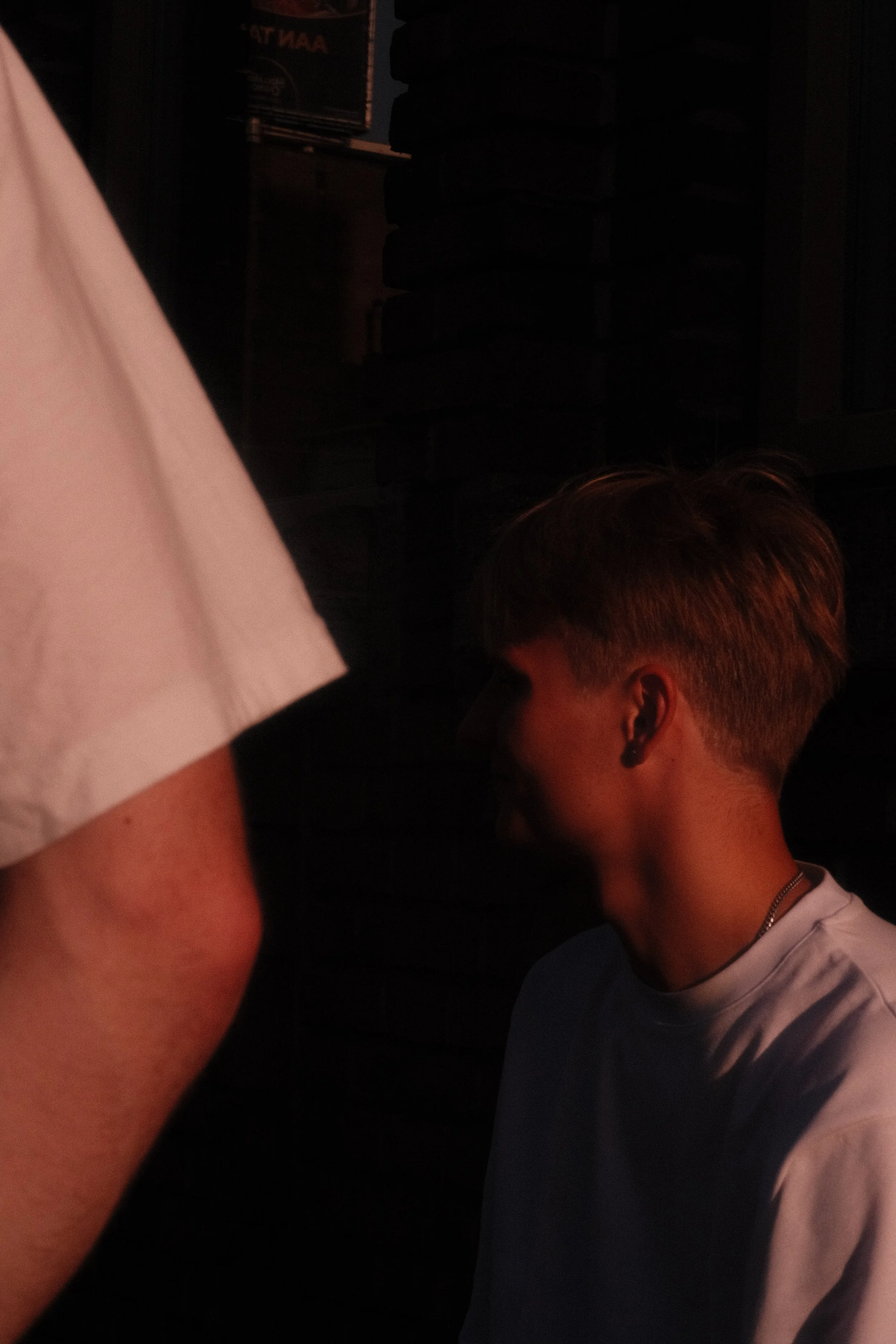 A young man with short, light brown hair and a nose piercing is sitting against a dark brick wall, illuminated by dim, reddish lighting.