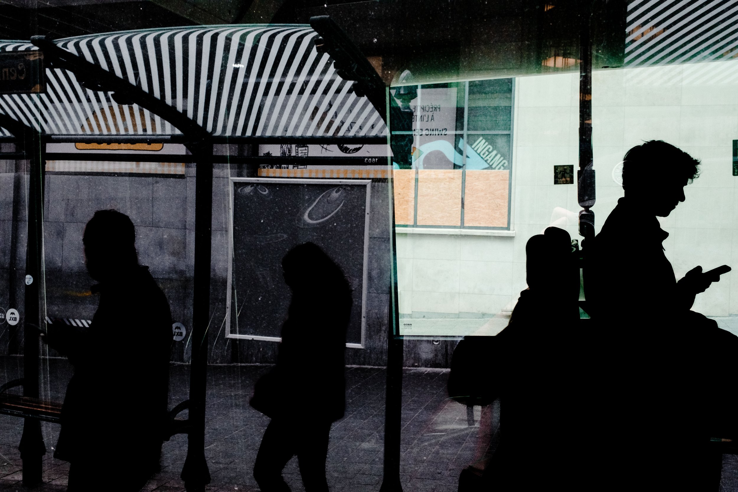 Silhouettes of people standing and using their phones inside a building, viewed through a glass window with reflections of the outside environment.
