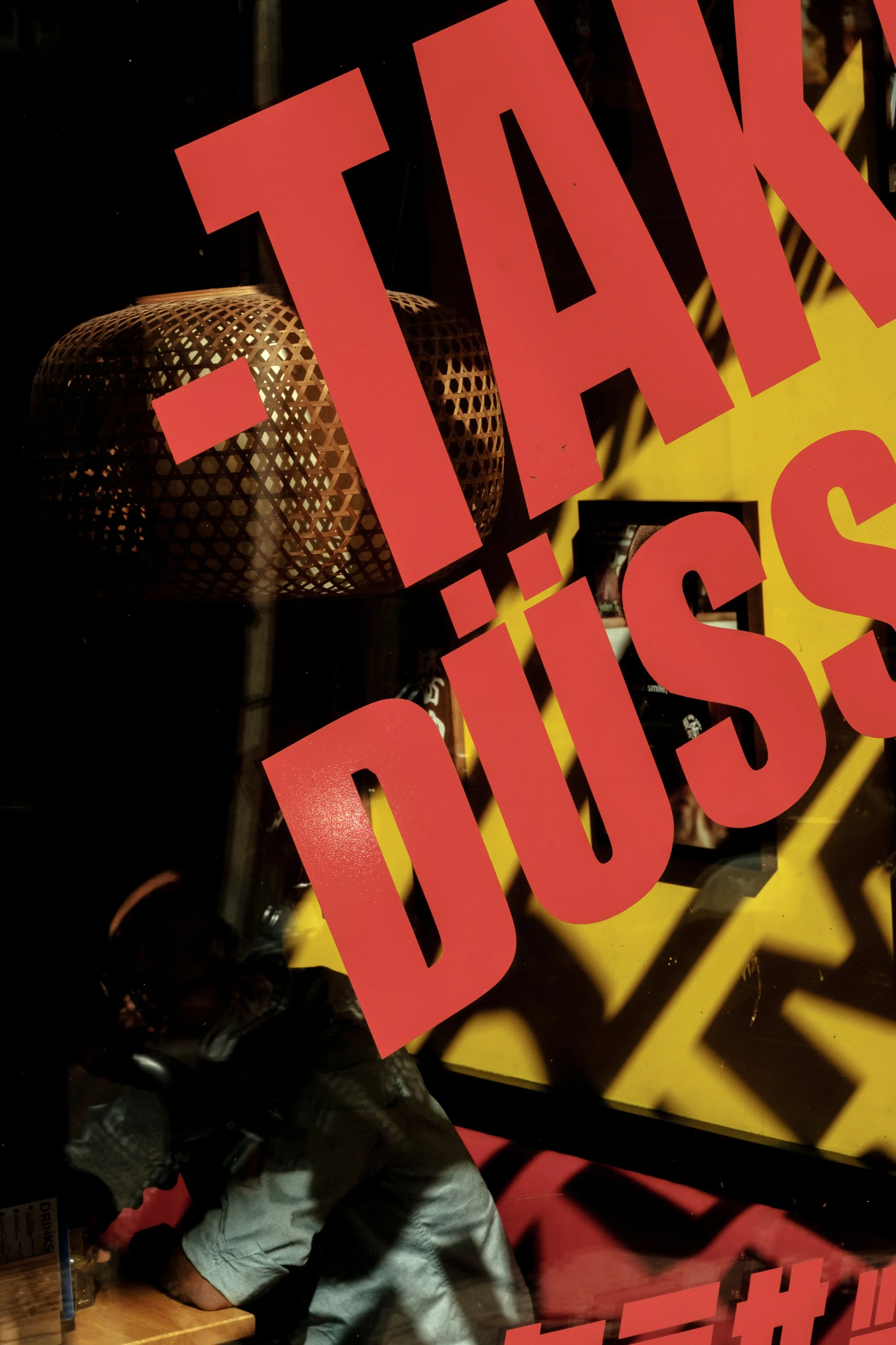 A storefront window with large red text that says 'TALK - DISS' partially visible, with a reflection of a woven pendant light and a person wearing a hoodie inside the shop.