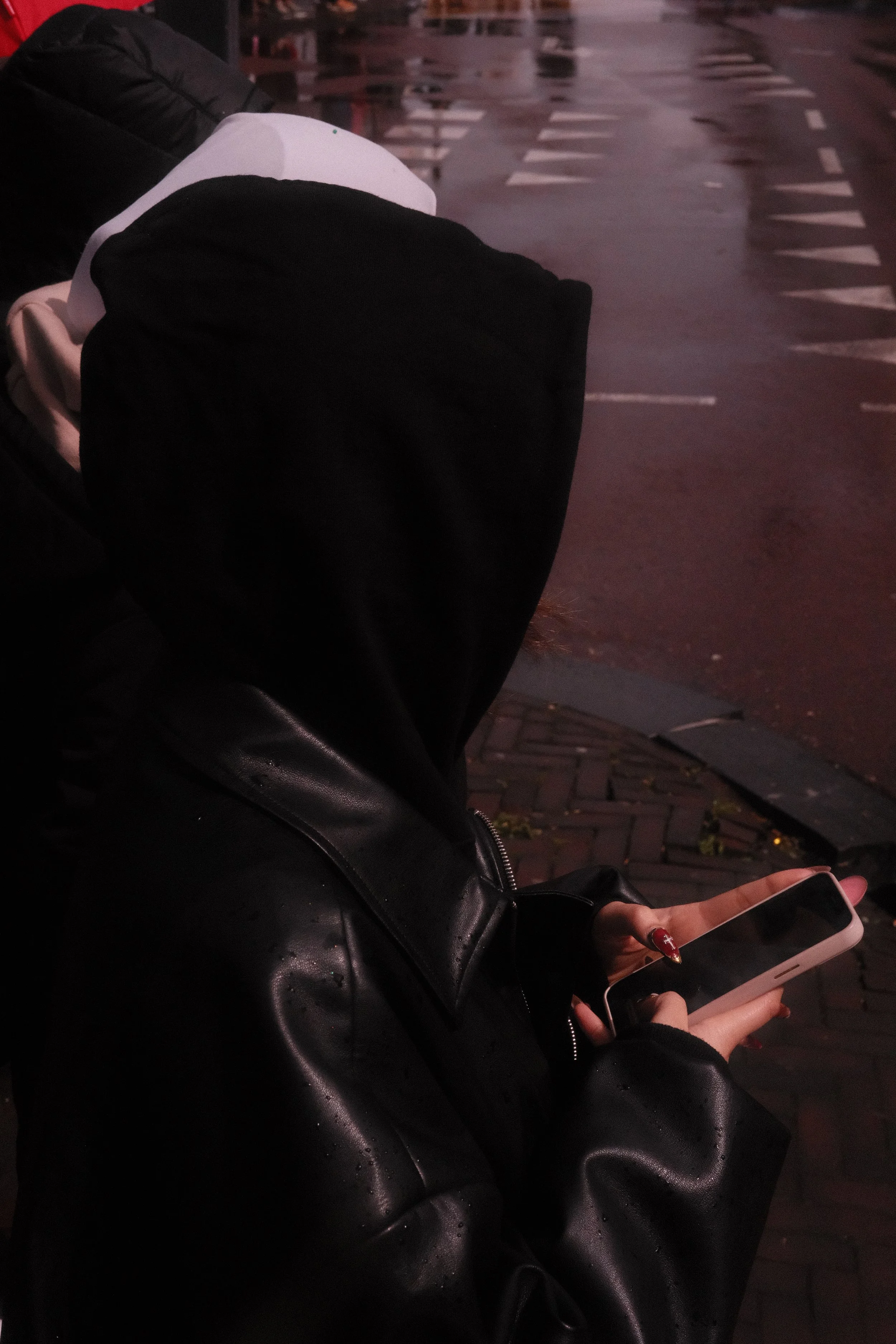 Person wearing a black hood and leather jacket sitting on a bench at a rainy street, holding a smartphone