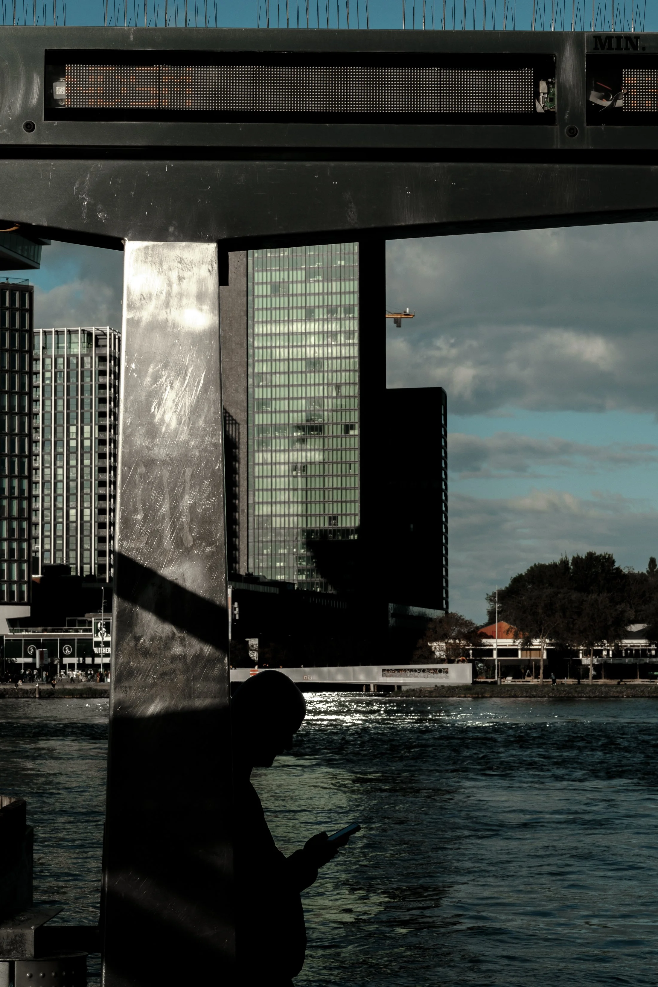 Silhouette of a person using a smartphone near a river with city buildings and a cloudy sky in the background.