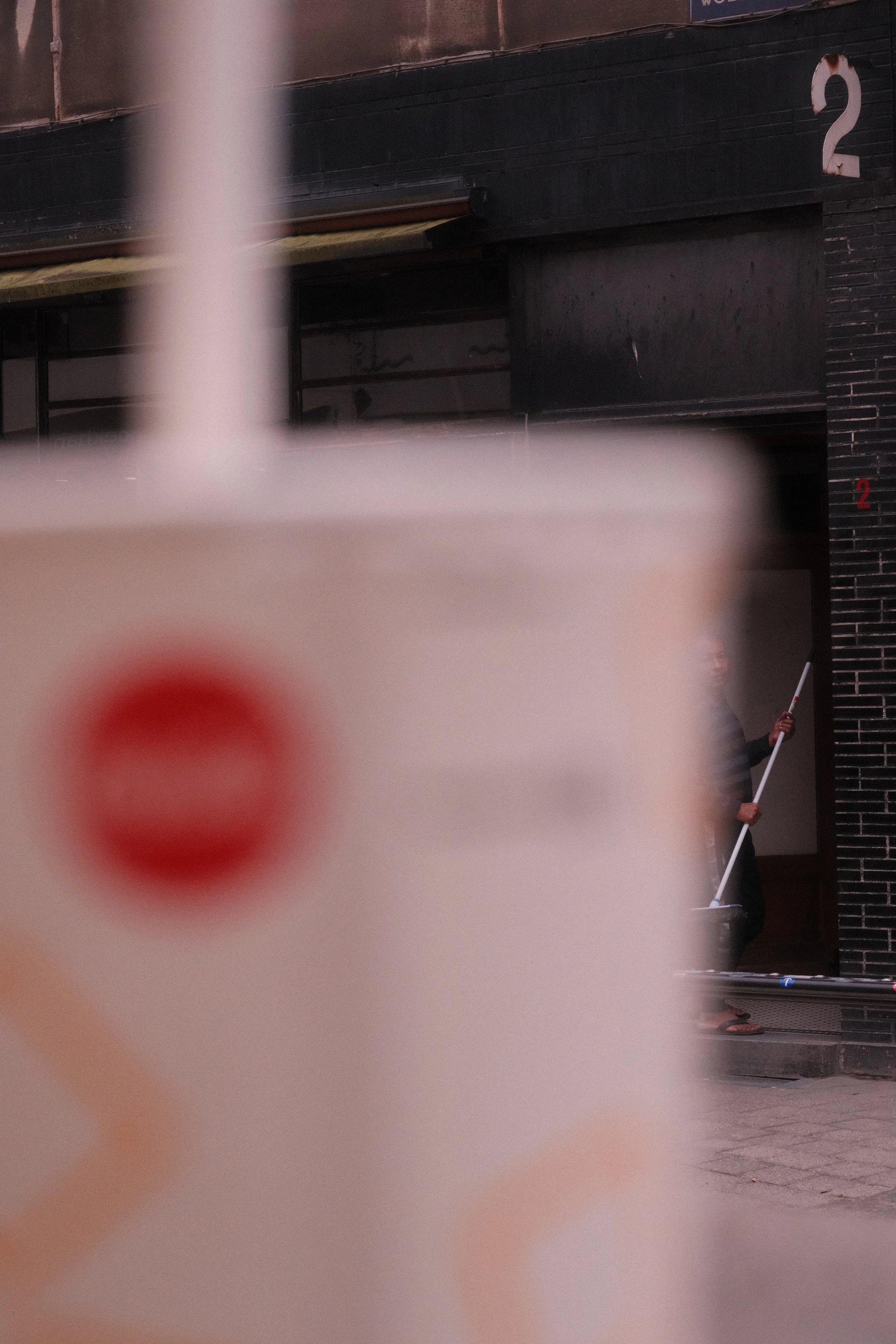 A person sweeping a sidewalk in front of a black brick building with the number 2 on it, partially obscured by a large, out-of-focus poster or sign in the foreground.