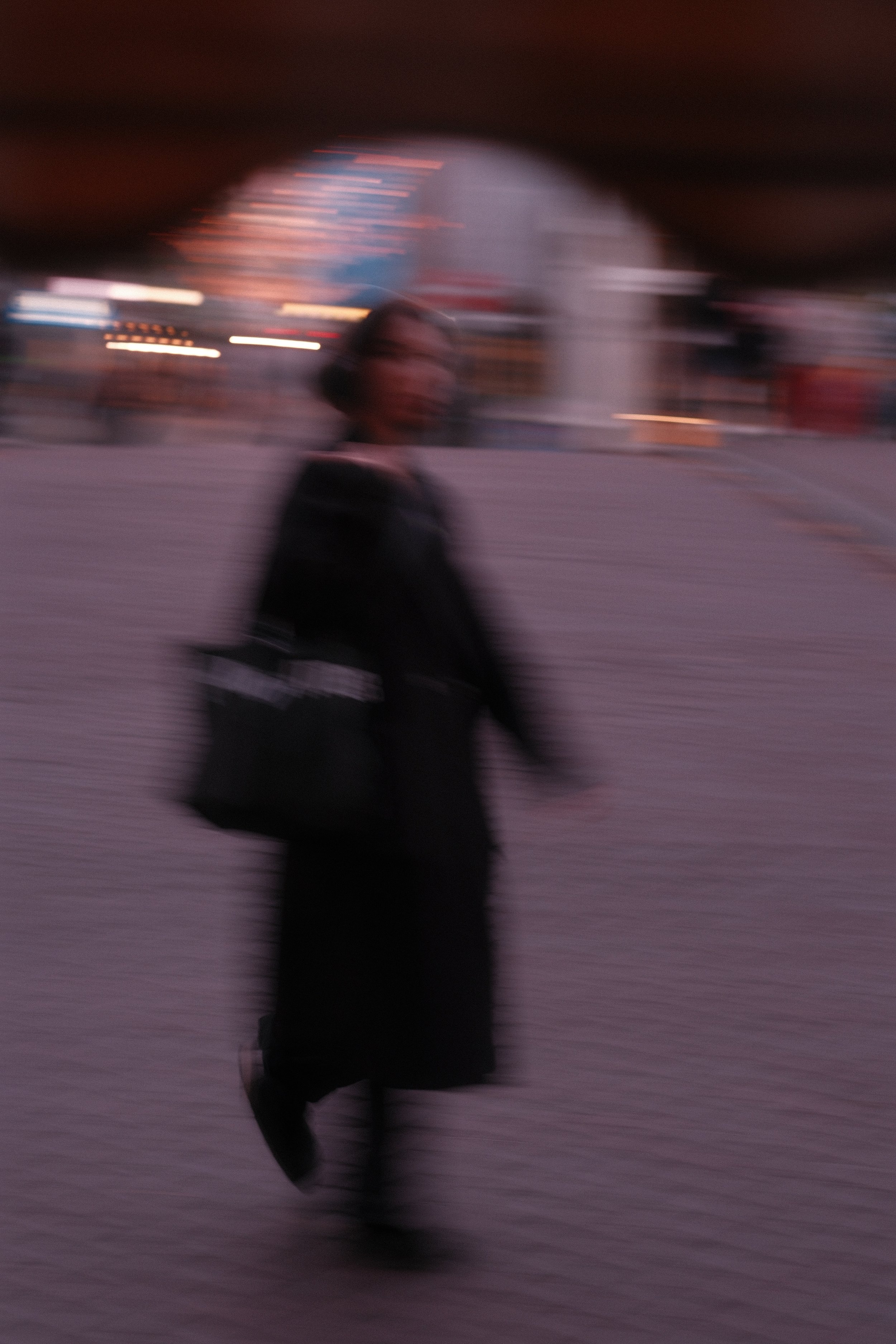 A blurred image of a person dressed in dark clothing walking outdoors, carrying a bag, with colorful lights and structures in the background.