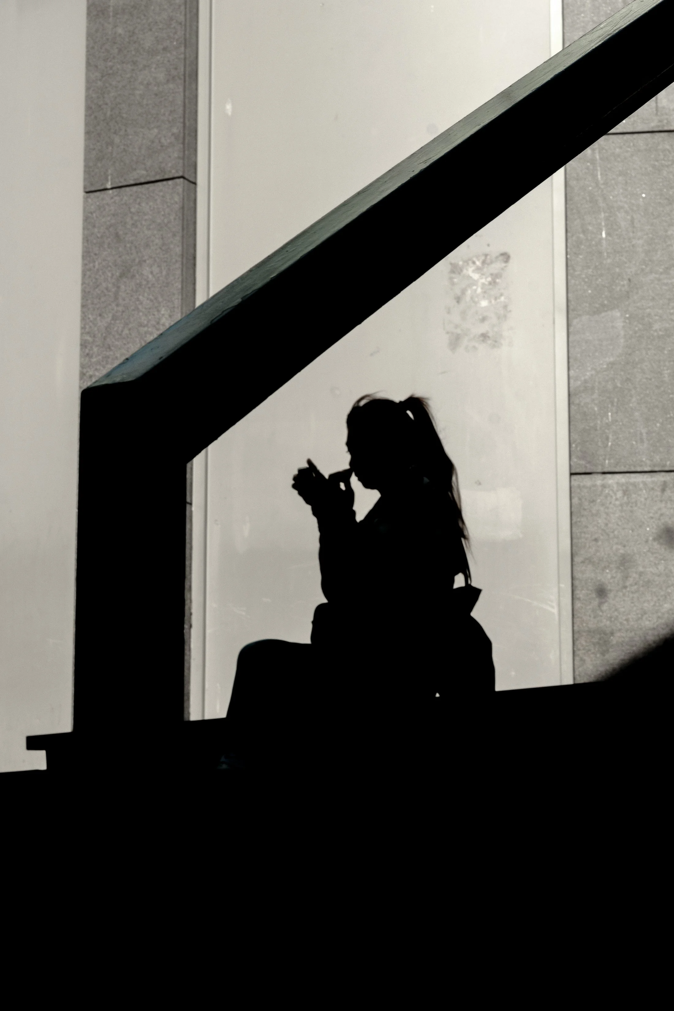 Silhouette of a girl sitting on stairs, holding a phone and taking a selfie or looking at the phone.