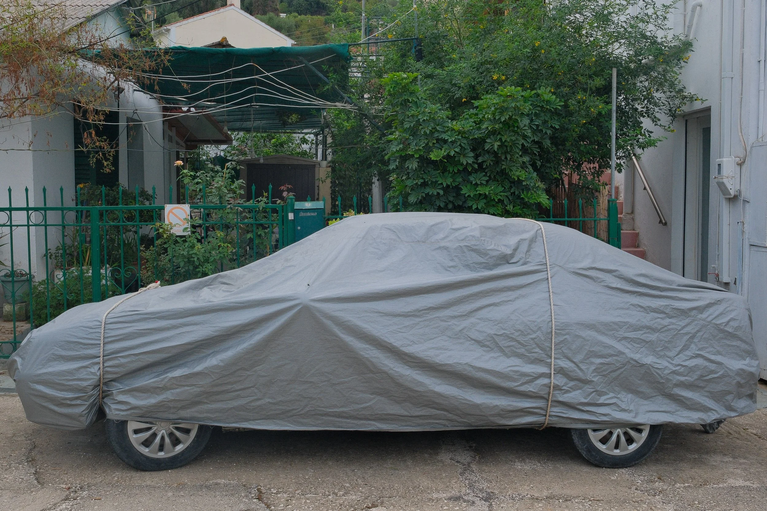 A car covered with a gray protective cover positioned on a gray concrete ground in front of a green fence and a residential area with lush trees and some staircases.