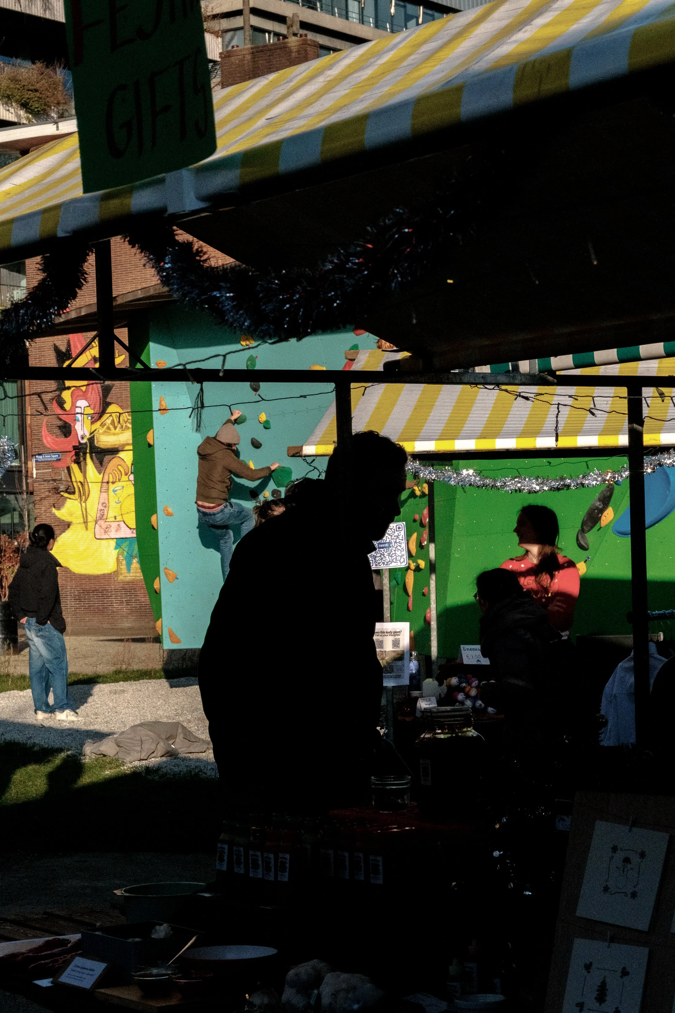 People at an outdoor night market, with a climbing wall in the background, decorated with holiday tinsel, and various vendor stalls at the foreground.