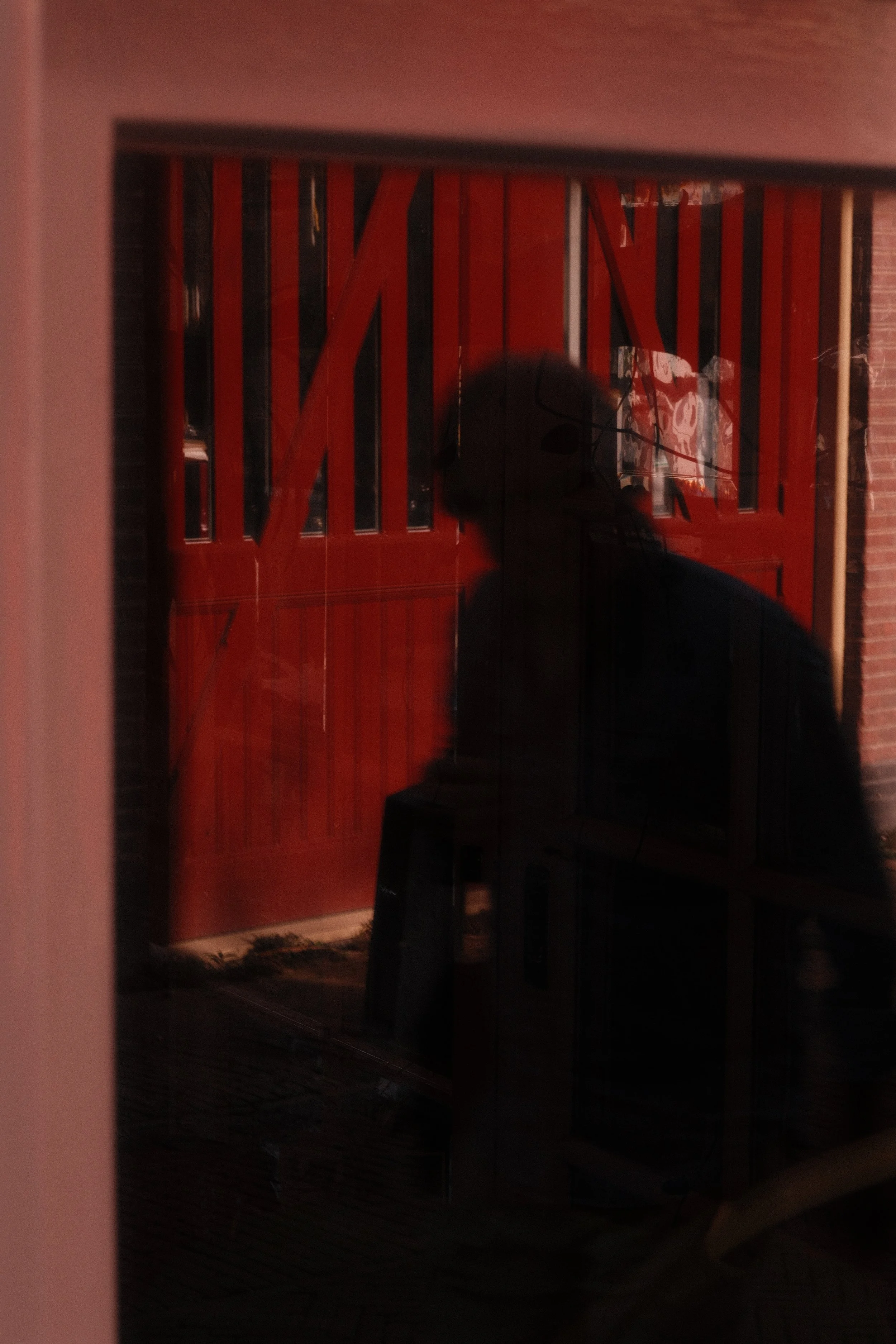 A reflection of a person with glasses, wearing a hat, seen on a glass surface, with a red wooden wall and a brick wall in the background.