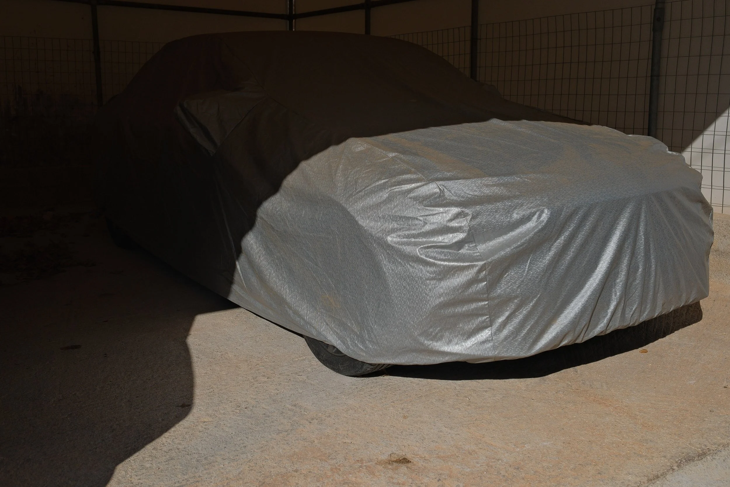 A car covered with a silver protective tarp parked on a concrete surface, shadow cast on the ground.