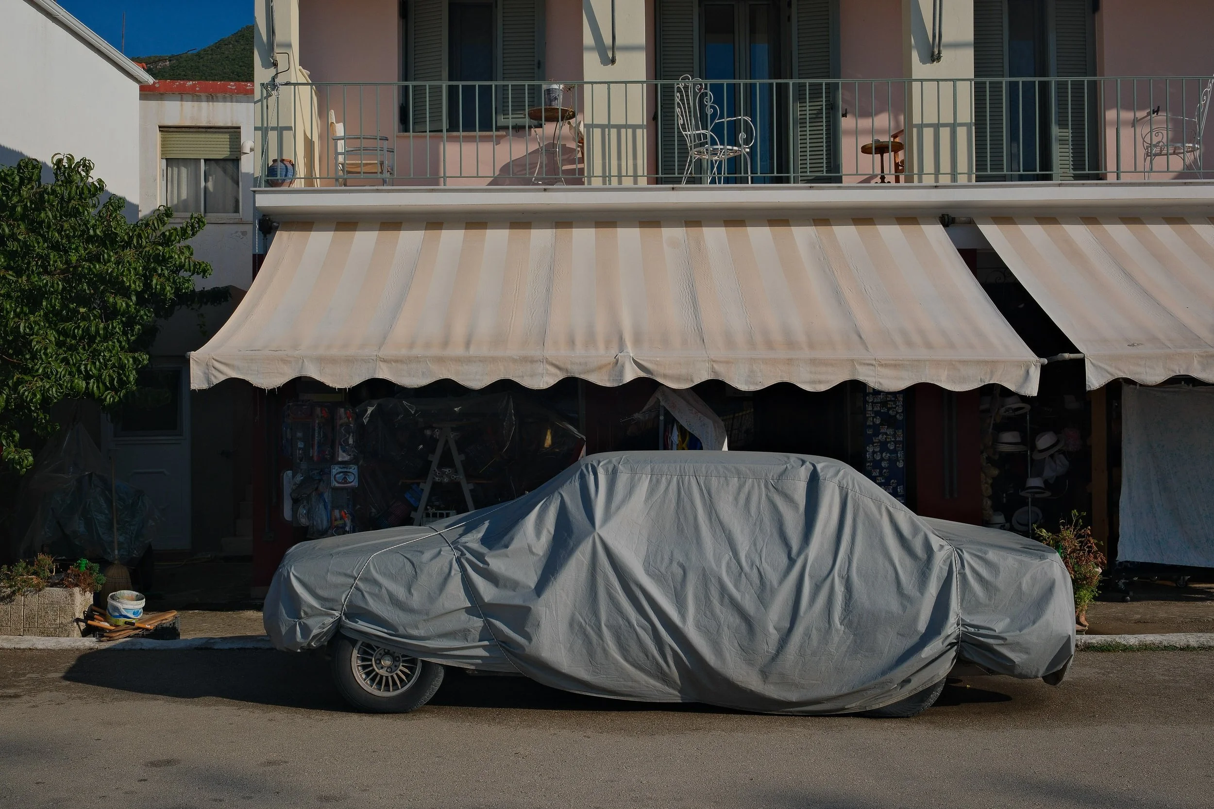 A car covered with a gray car cover parked in front of a building with a beige and white striped awning, on a street during daylight.
