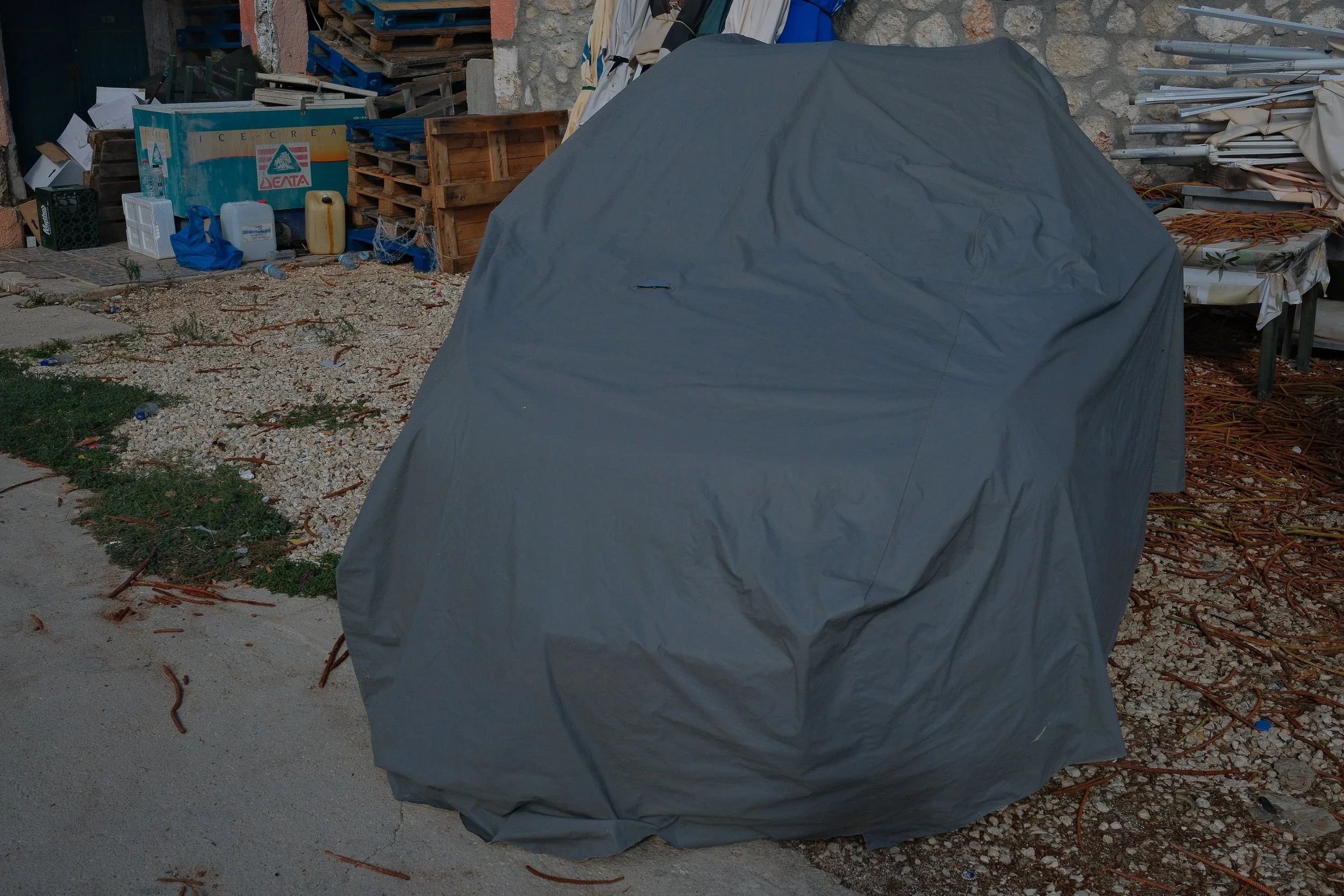 A large object covered with a gray tarp in an outdoor area with gravel ground, surrounded by wooden pallets, containers, a table, and cluttered items.