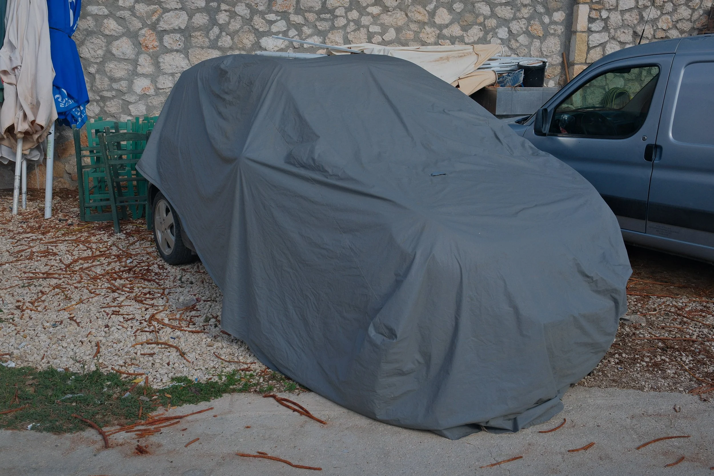 A car covered with a gray car cover parked next to a second car on a gravel surface, with a stone wall in the background. There are outdoor chairs, umbrellas, and miscellaneous items nearby.