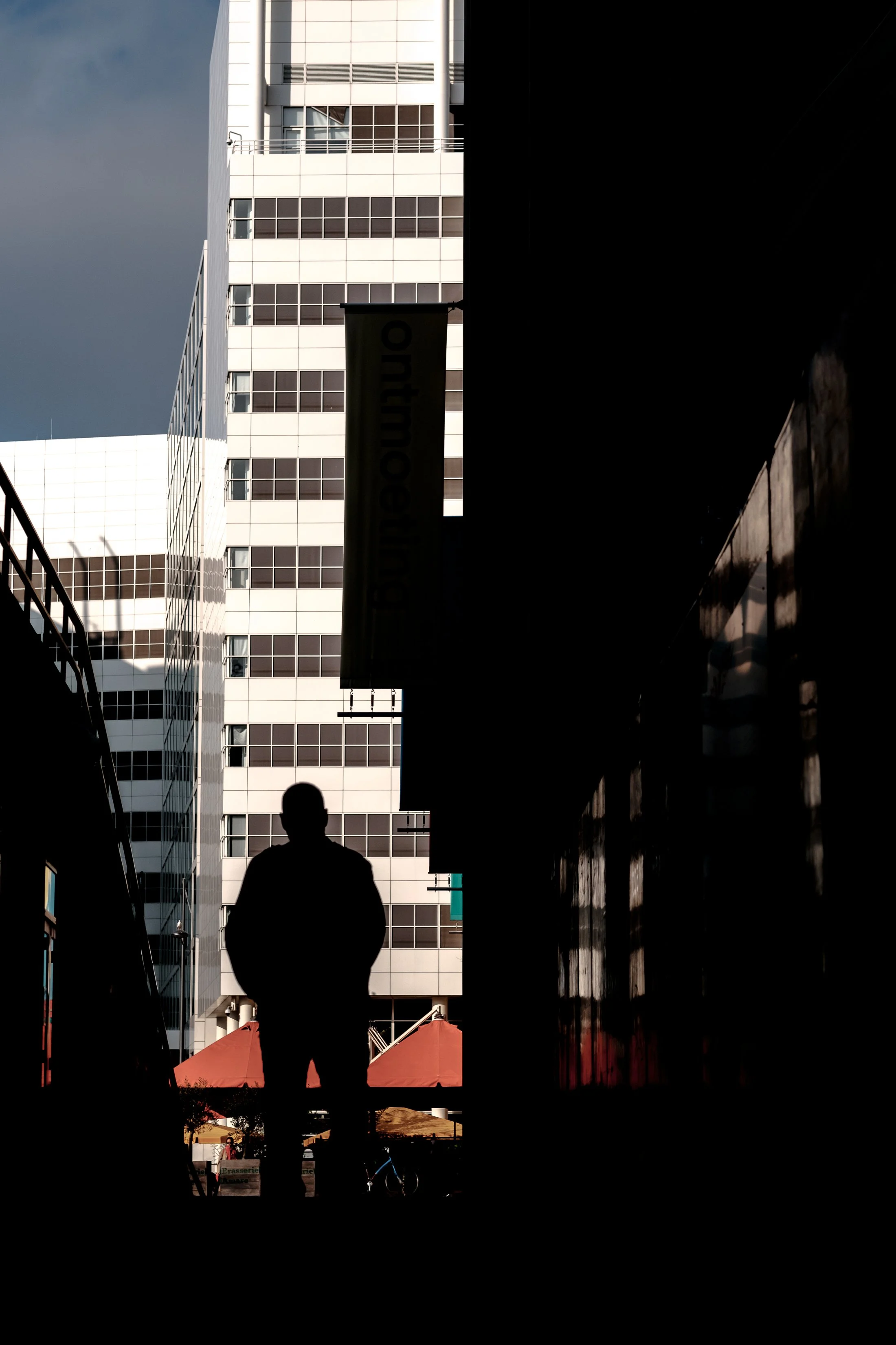 A person in silhouette standing in an alleyway with tall modern building in the background, some tents with red tops are visible at the bottom, and a bicycle is also present.