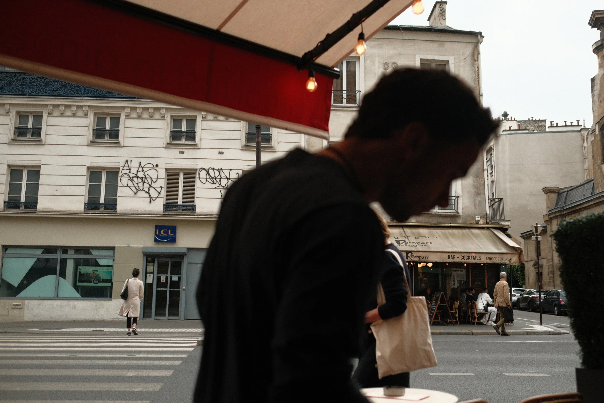 A silhouette of a man inside a cafe or restaurant, with an urban street scene outside including pedestrians, buildings, and a bar with outdoor seating.