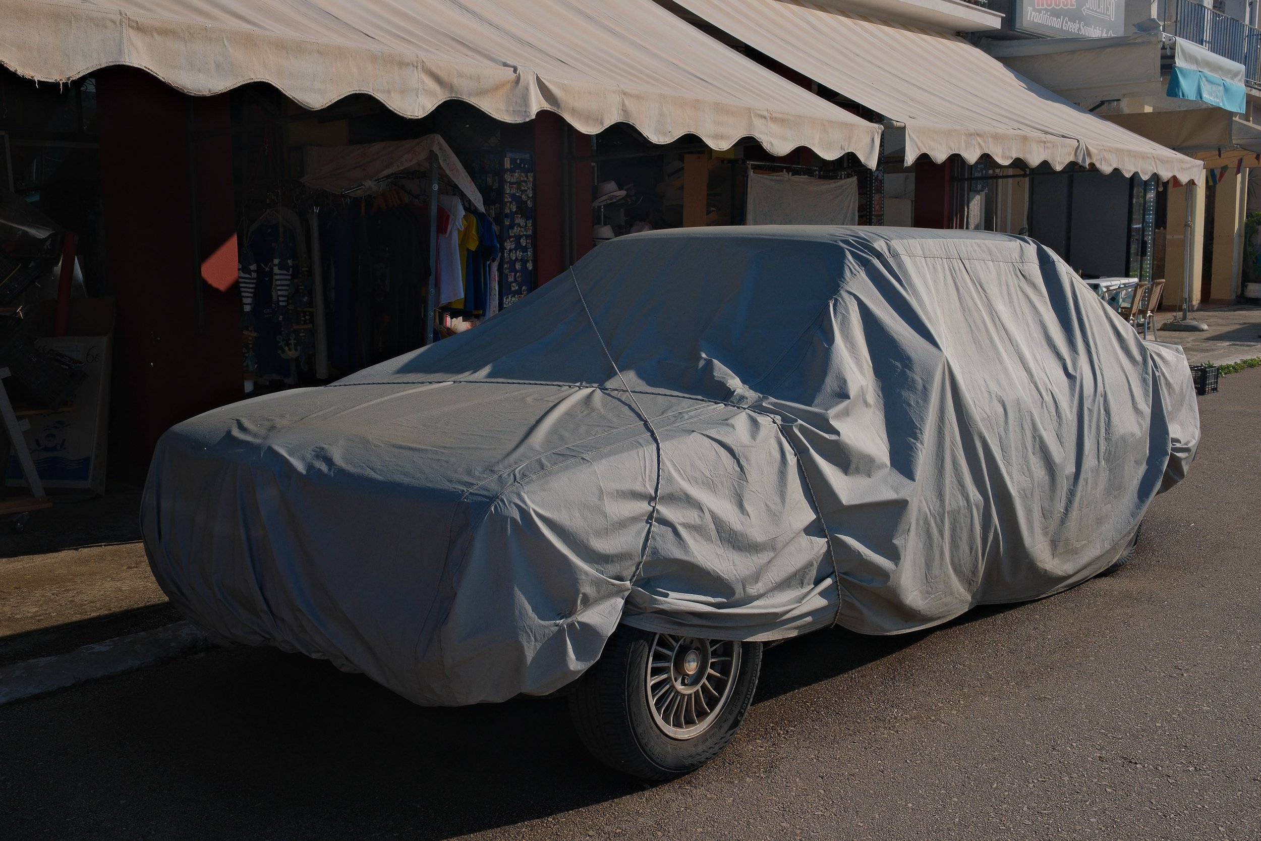 A car covered with a gray car cover parked on the street in front of a storefront with an awning and clothing and accessories inside.