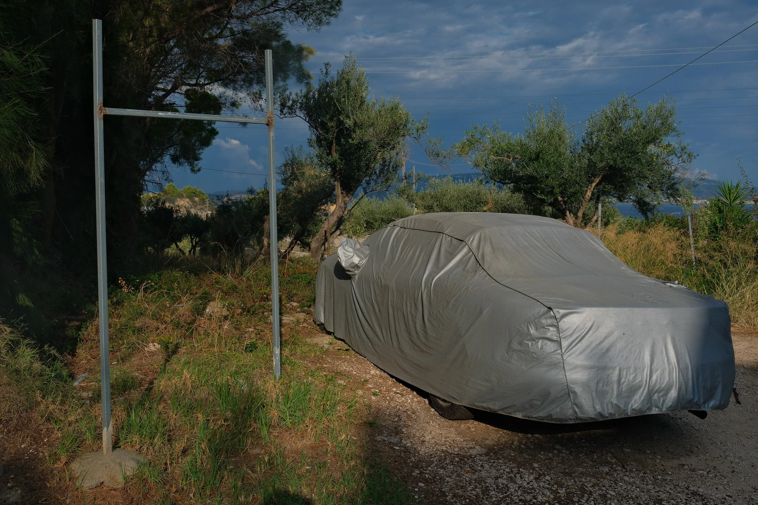 A car covered with a gray car cover parked on a dirt path, surrounded by trees and shrubs, with a partially cloudy sky in the background.