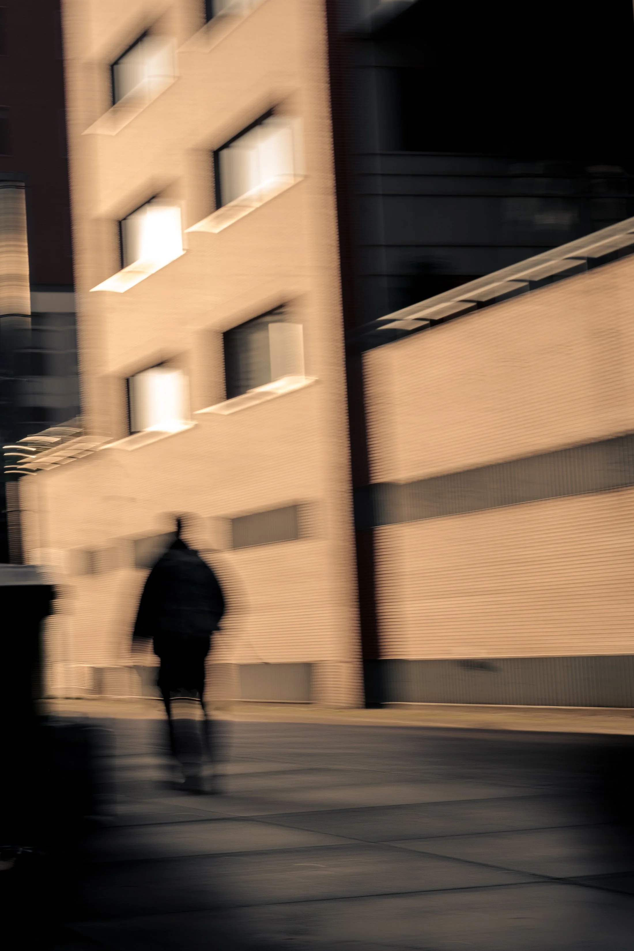 A blurred city street scene with a person walking past a beige brick building.