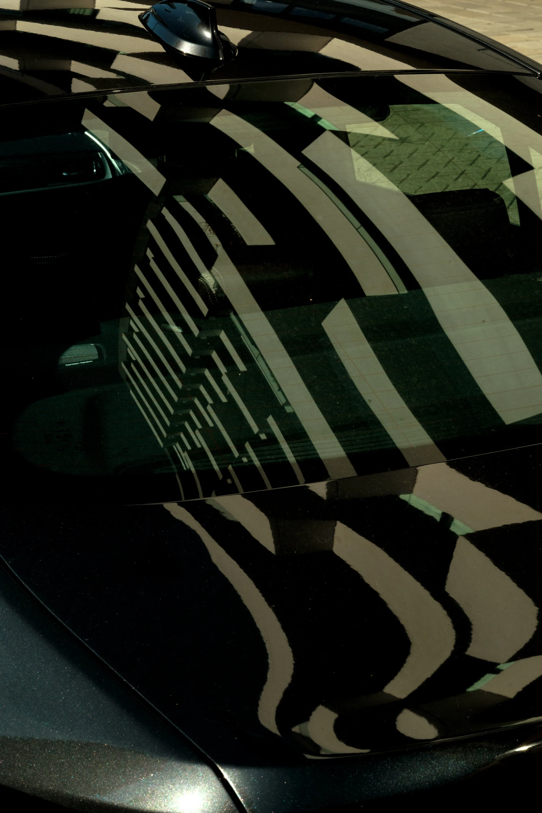 Close-up of the roof and windshield of a black car, reflecting a building's striped architectural pattern on the glass.