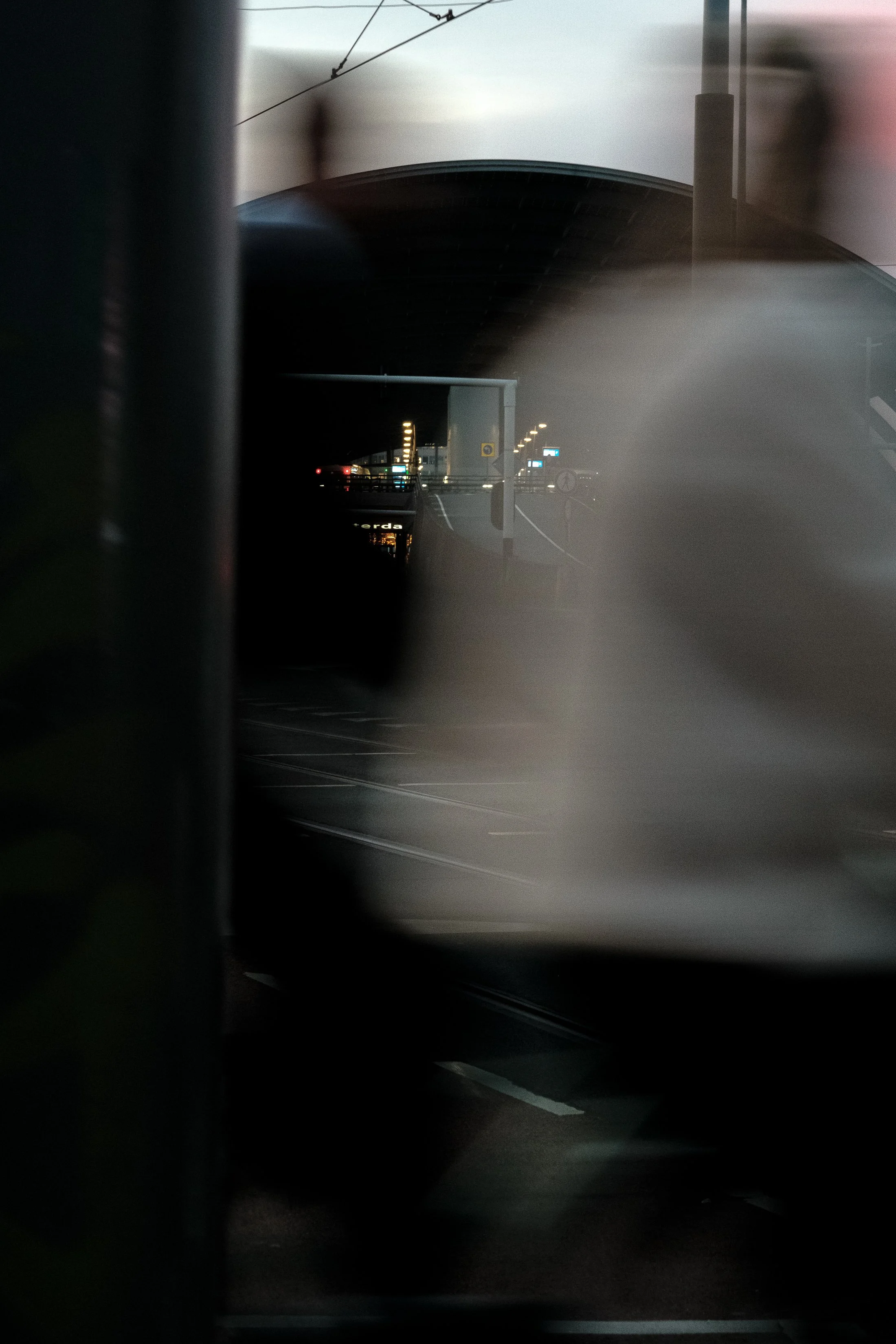 Blurred image of a train station seen through a window or reflection, with tracks and illuminated signs in the background, possibly during dusk or nighttime.