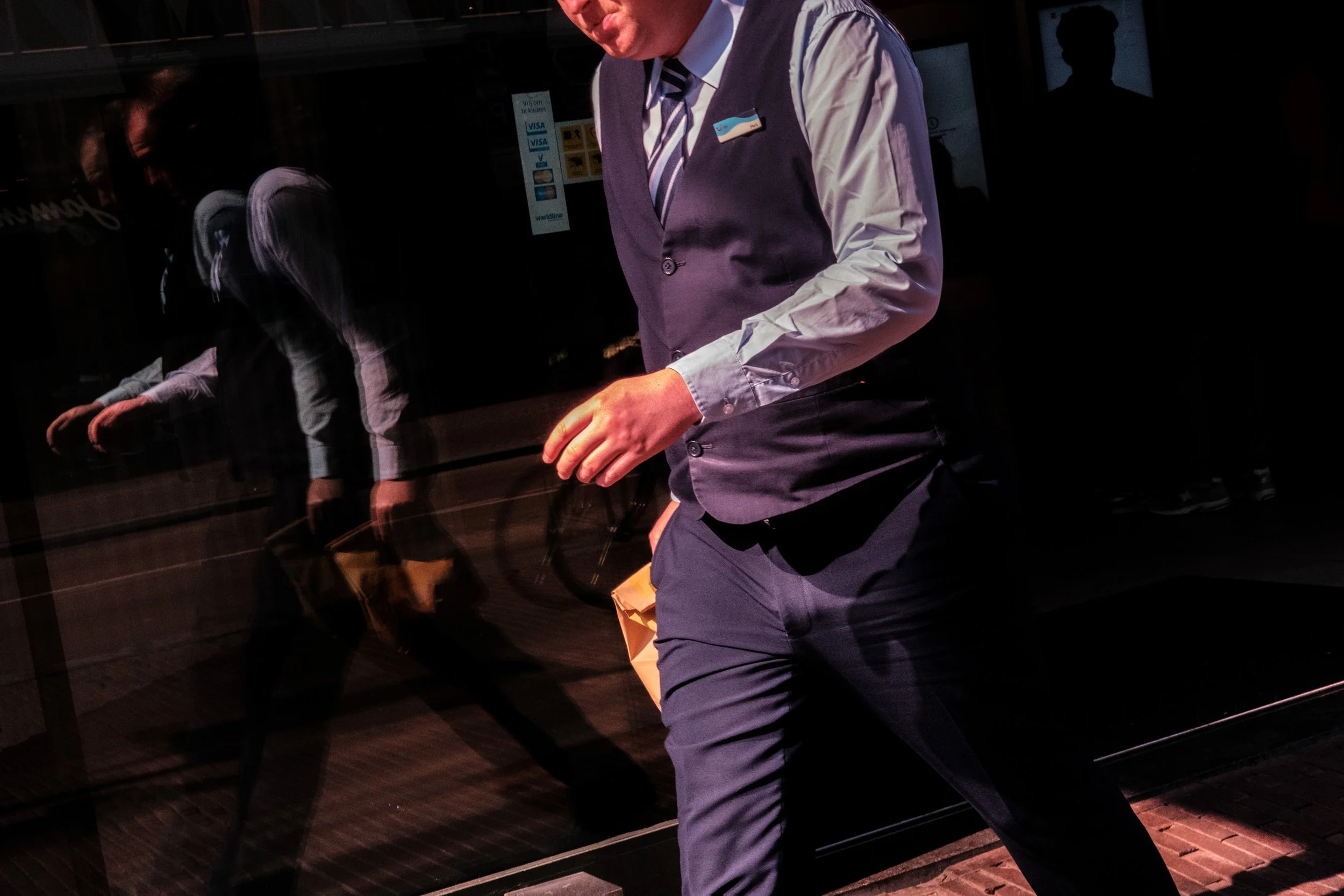 A man wearing a vest and dress shirt walking past a glass building with his reflection visible on the glass.