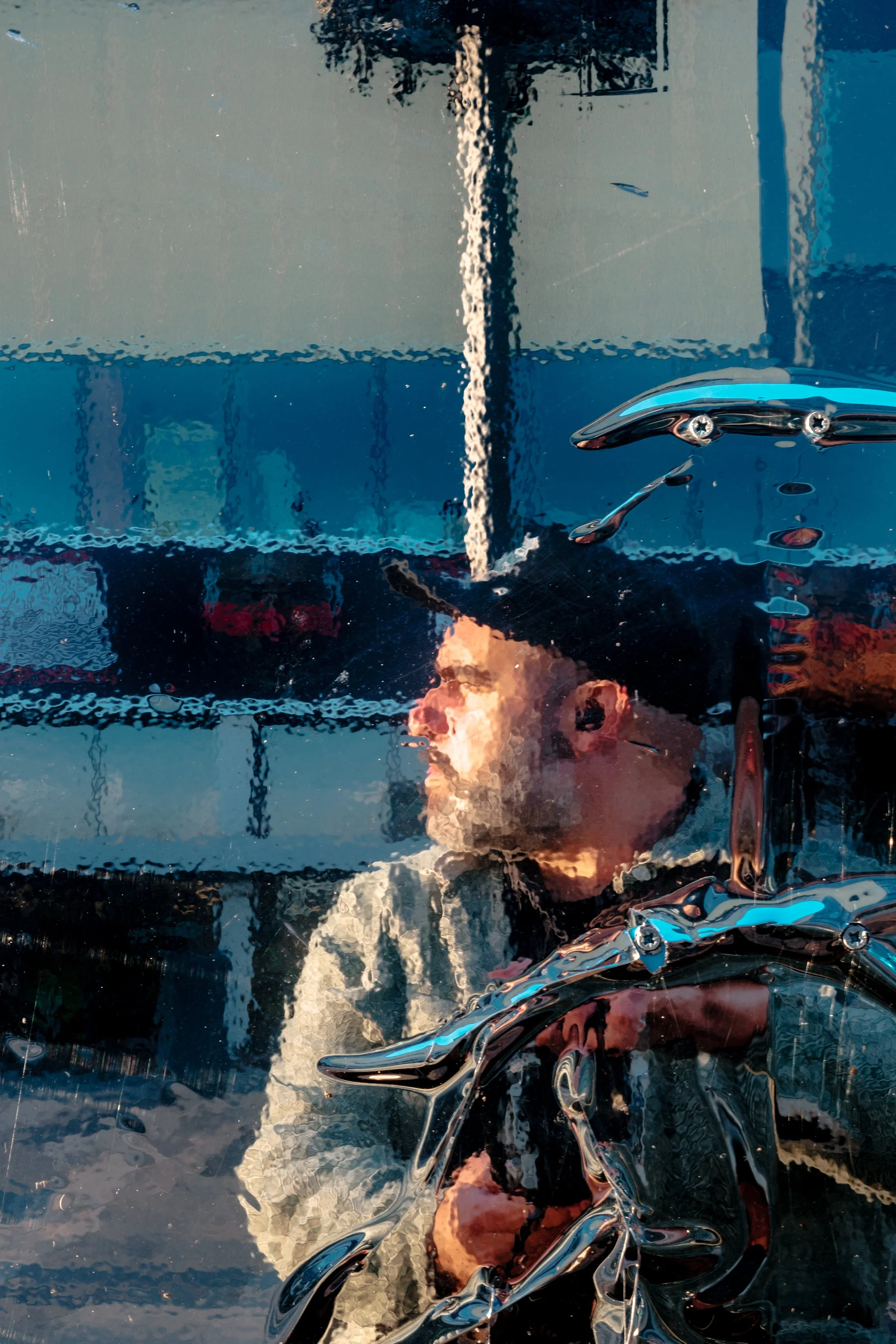 The reflection of a man wearing sunglasses and a jacket is seen on a reflective surface, possibly a glass or a metallic object, with distorted reflections and light effects.