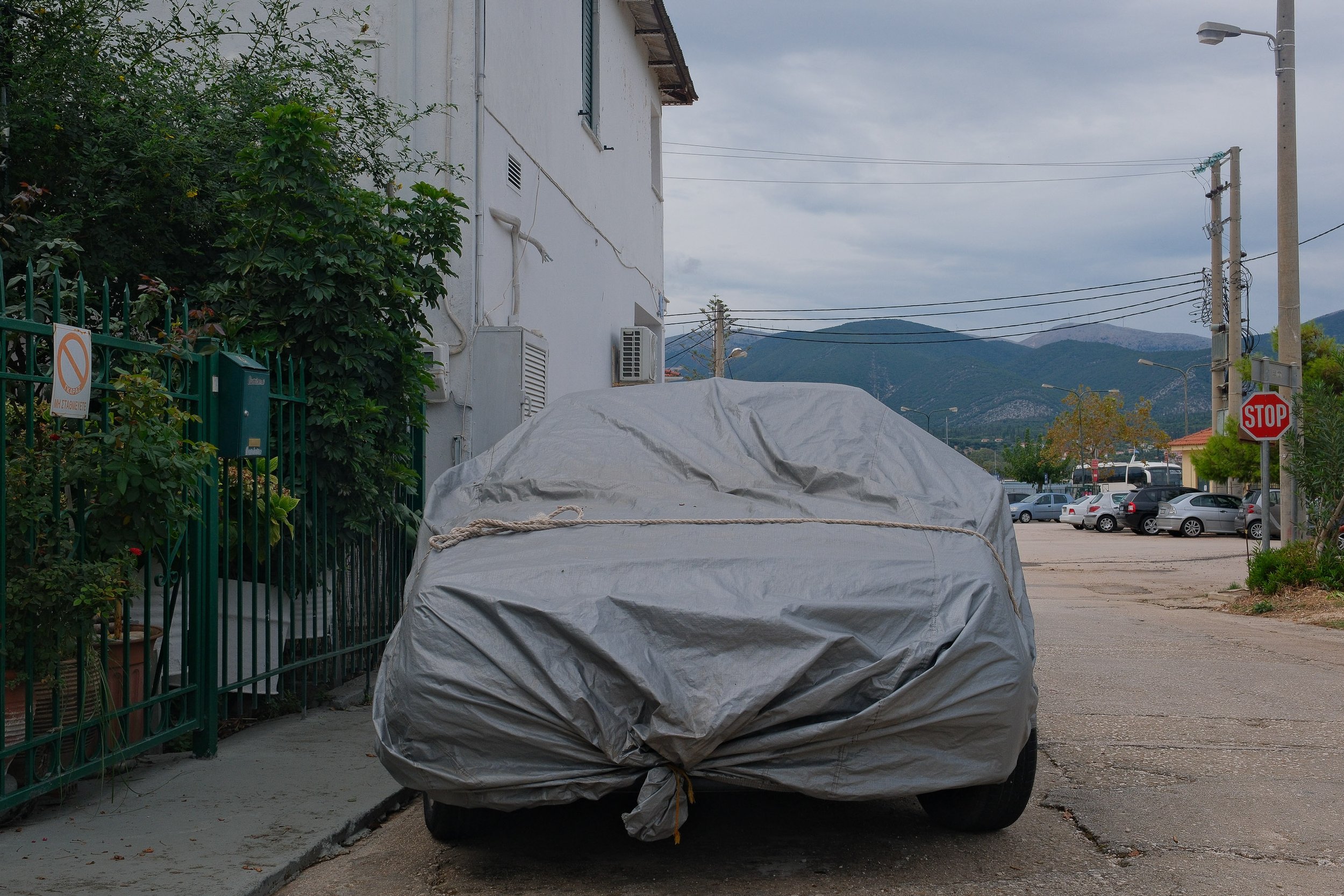 A car covered with a gray car cover secured with a rope, parked on a street near a green fence and some plants. The background shows mountains, cloudy sky, and other parked cars.