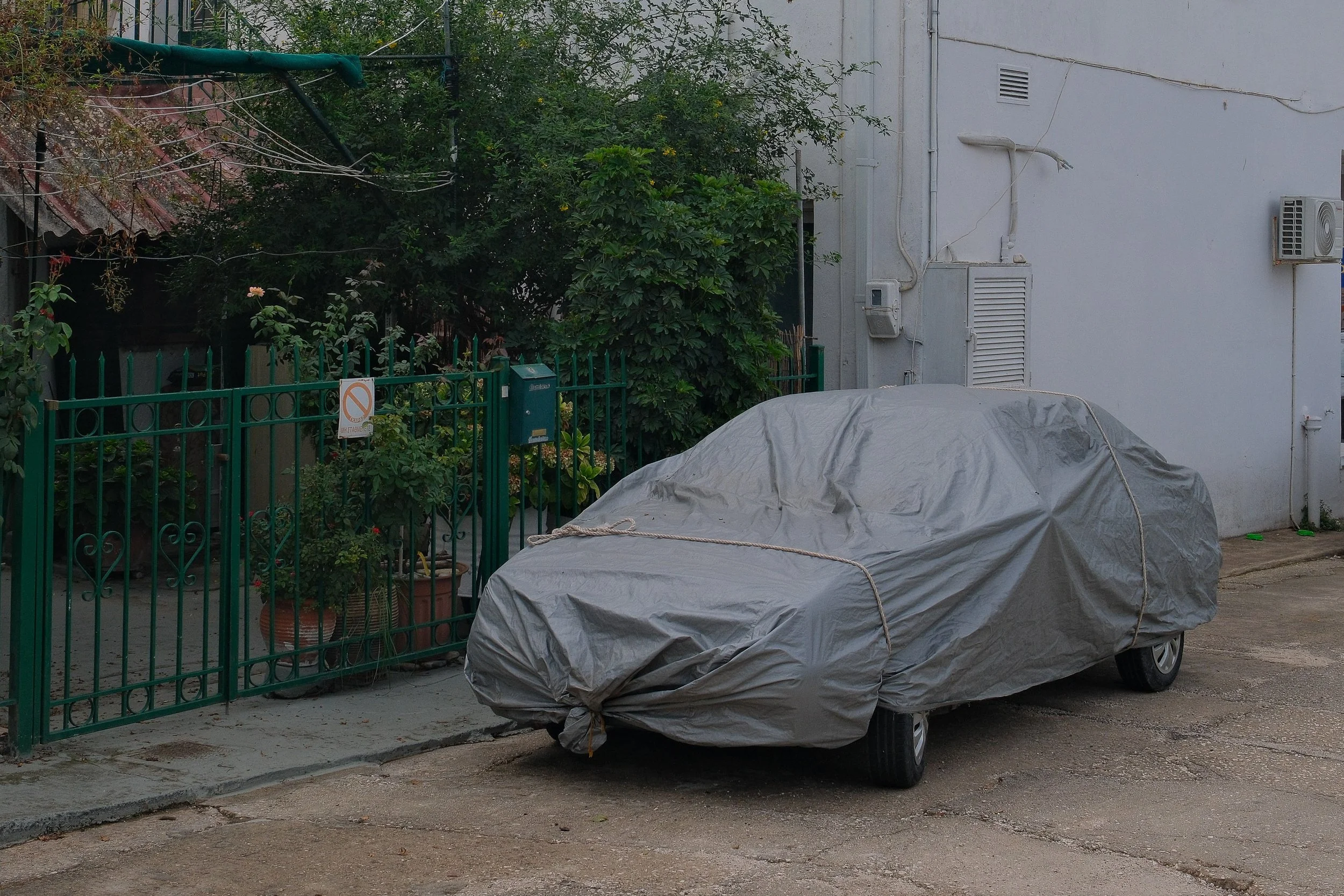 A car covered with a gray tarp parked on a street next to a green metal fence and a residential building.