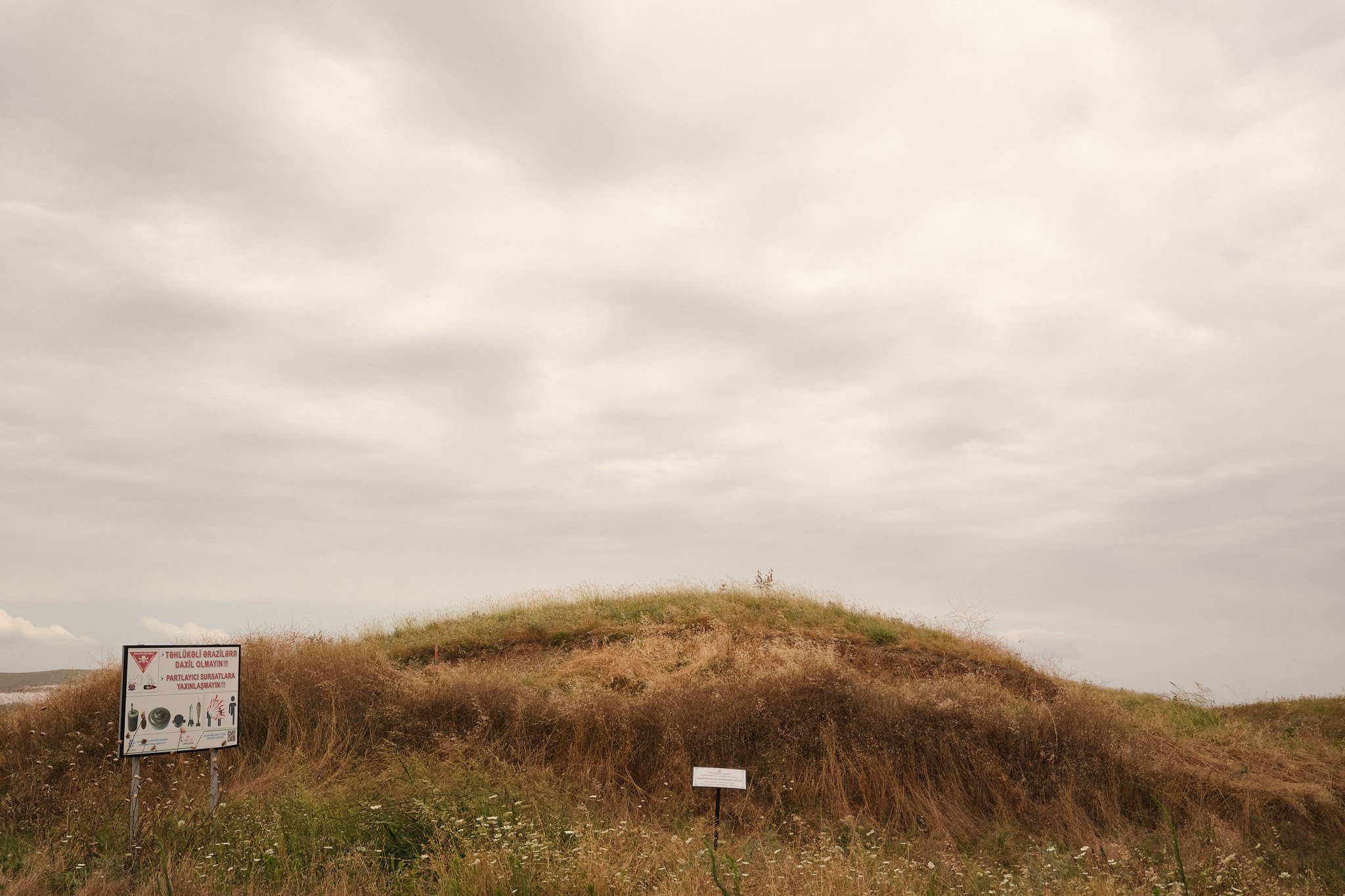 Signs warn of landmines and other unexploded weapons: “Do not enter dangerous areas. Do not approach explosive ordnance.” Aslan explained that these unassuming mounds were used as lookouts during the conflict; Nagorno-Karabakh, July 2026.