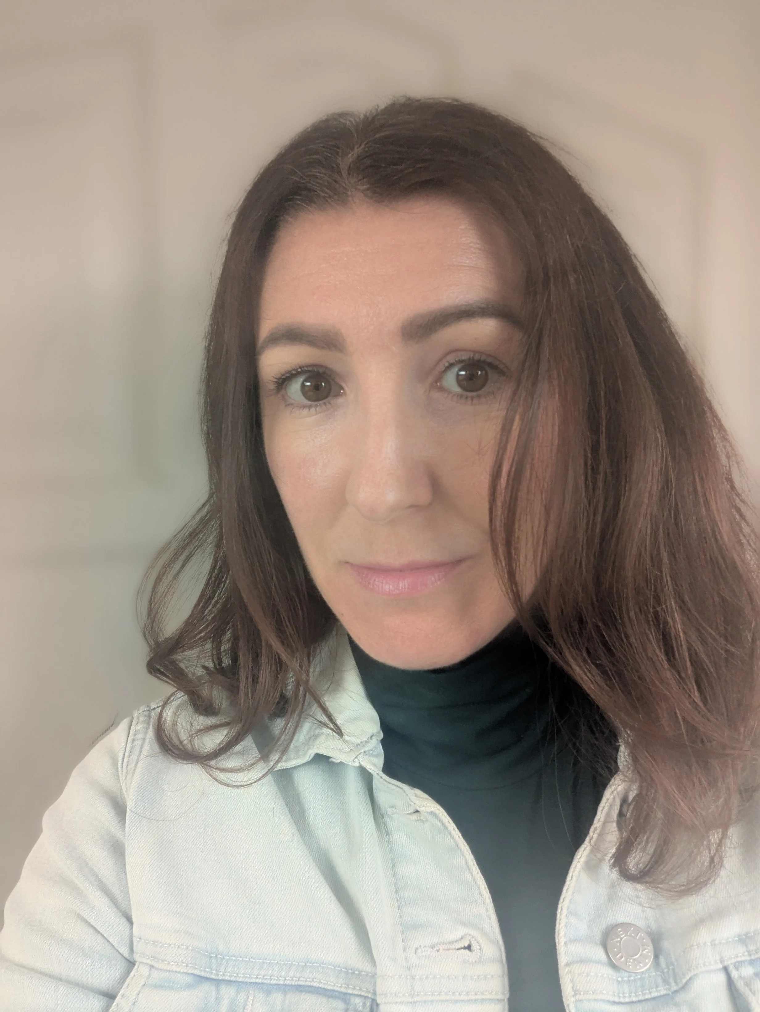 A woman with shoulder-length brown hair, hazel eyes, wearing a black turtleneck and a light-colored jacket, posing for a selfie indoors.