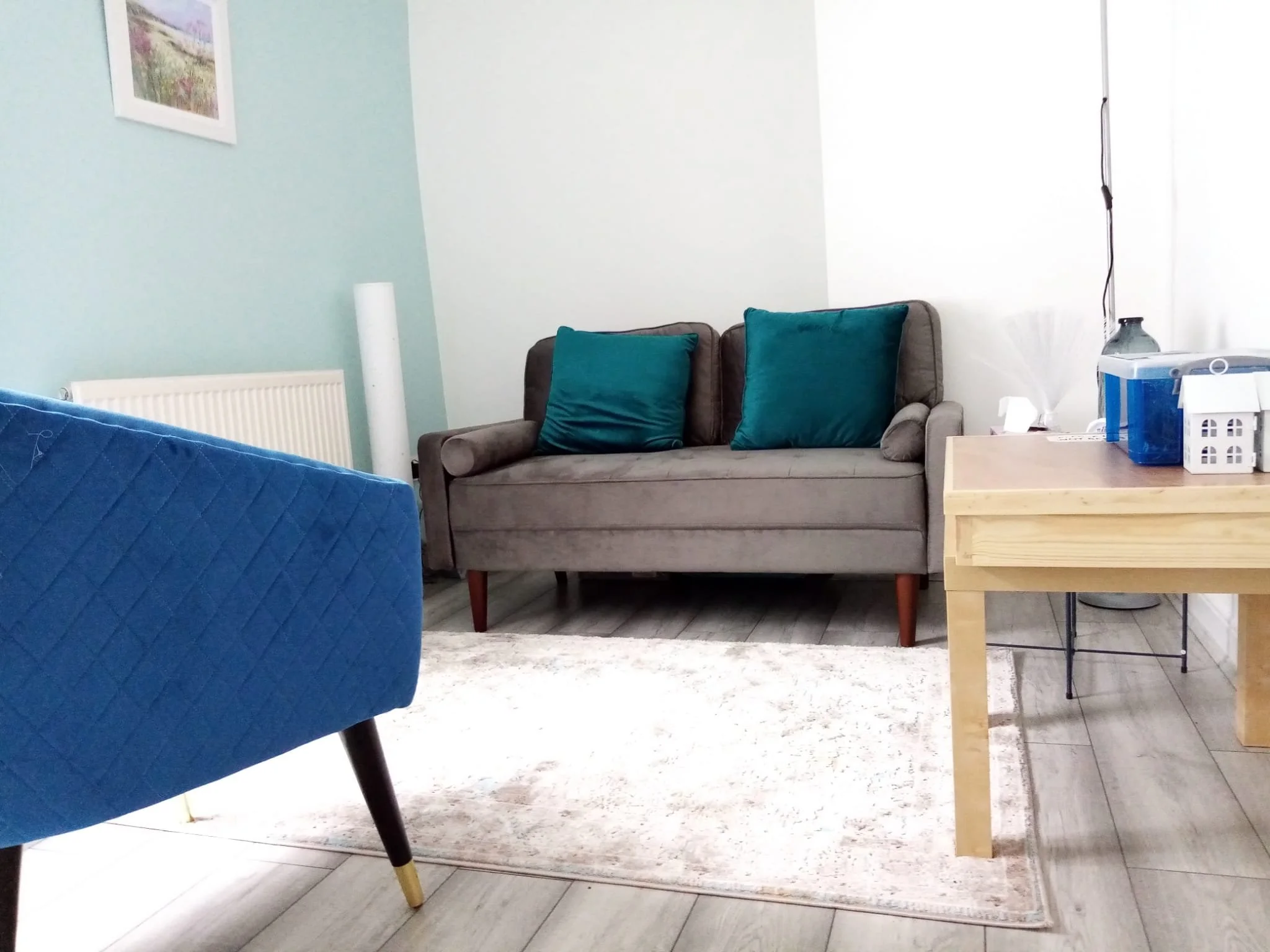 Living room with a gray sofa with teal pillows, a blue armchair, a wooden table with various items, a small white rug, and light-colored wood flooring.