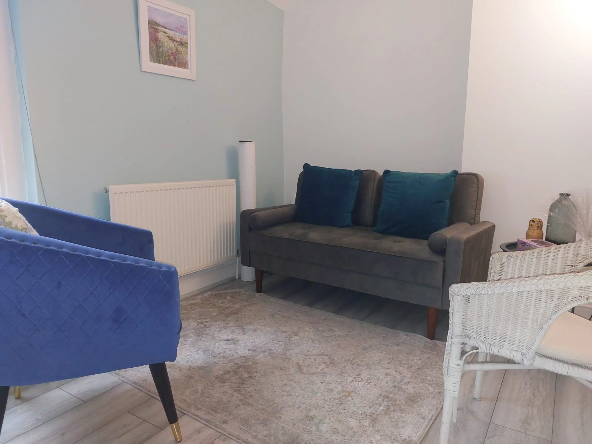 A cozy living room with a light blue wall, a picture frame with floral art, a white radiator, a grey sofa with two blue cushions, a white wicker chair, and a beige rug.