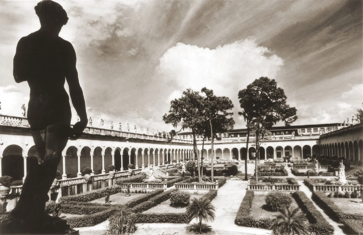 A black and white photograph of a courtyard with a large statue of a standing man in the foreground, surrounded by trees, neatly trimmed bushes, and classical architecture with arched walkways and balustrades.