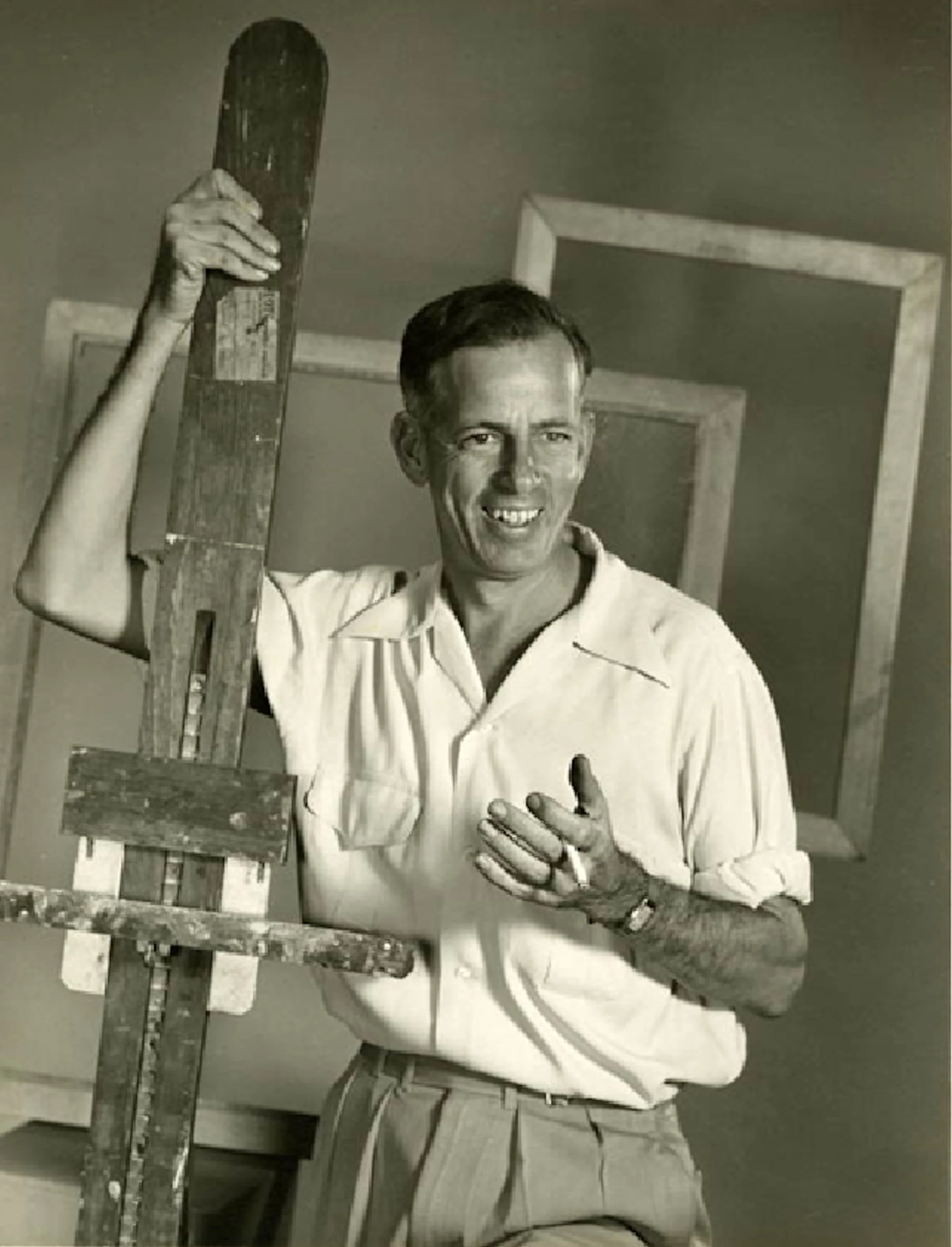 A smiling man holding an artist's palette and standing next to an easel, in front of empty picture frames, in a studio setting.