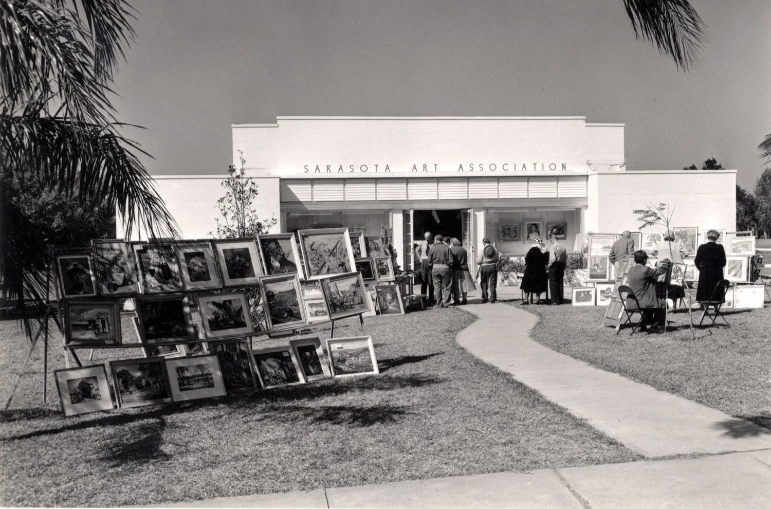 An outdoor art exhibition in front of the Sarasota Art Association building, displaying numerous framed paintings and artworks with several people viewing the art.