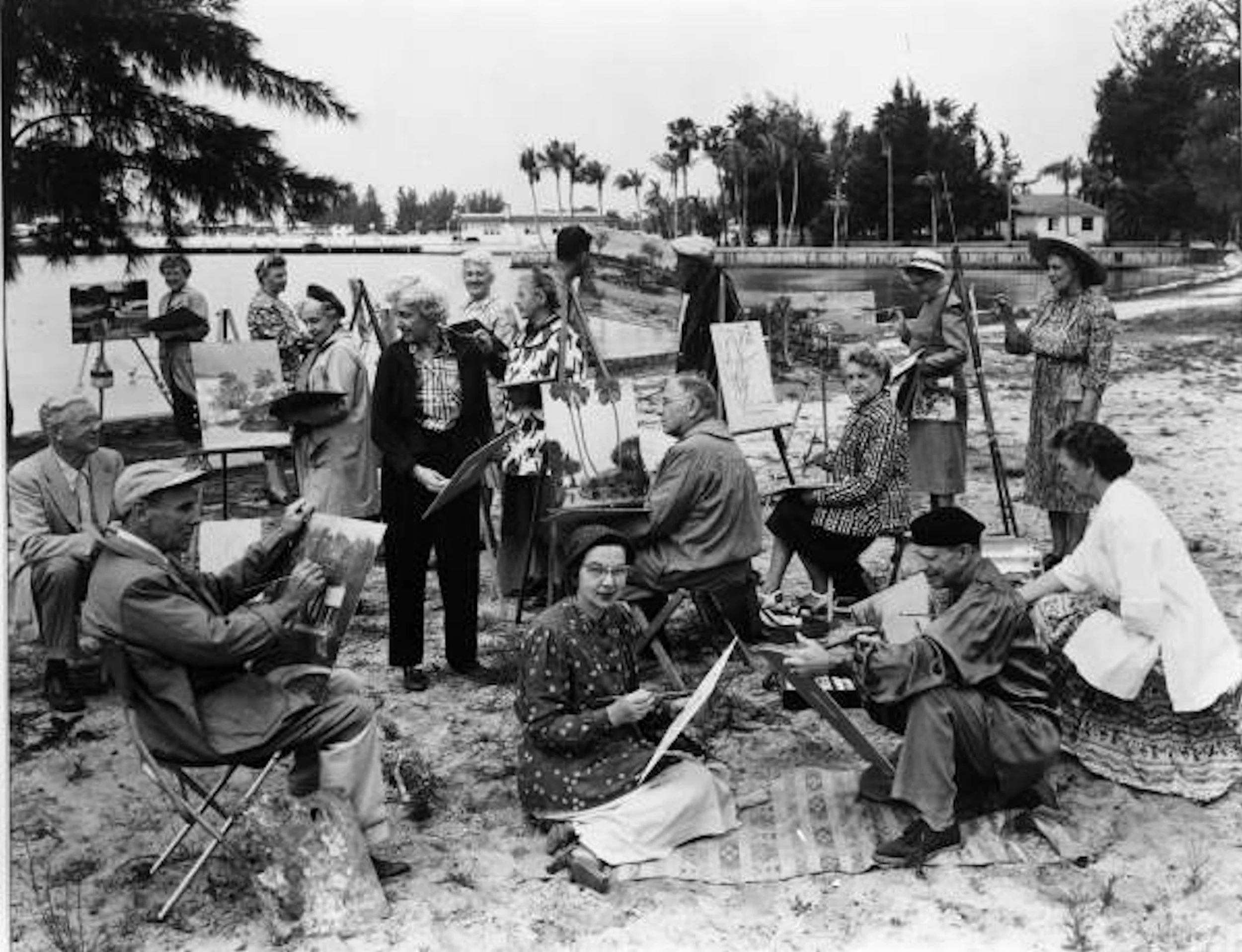 Group of people, mostly women, painting and drawing outdoors near a body of water, with trees and houses in the background.