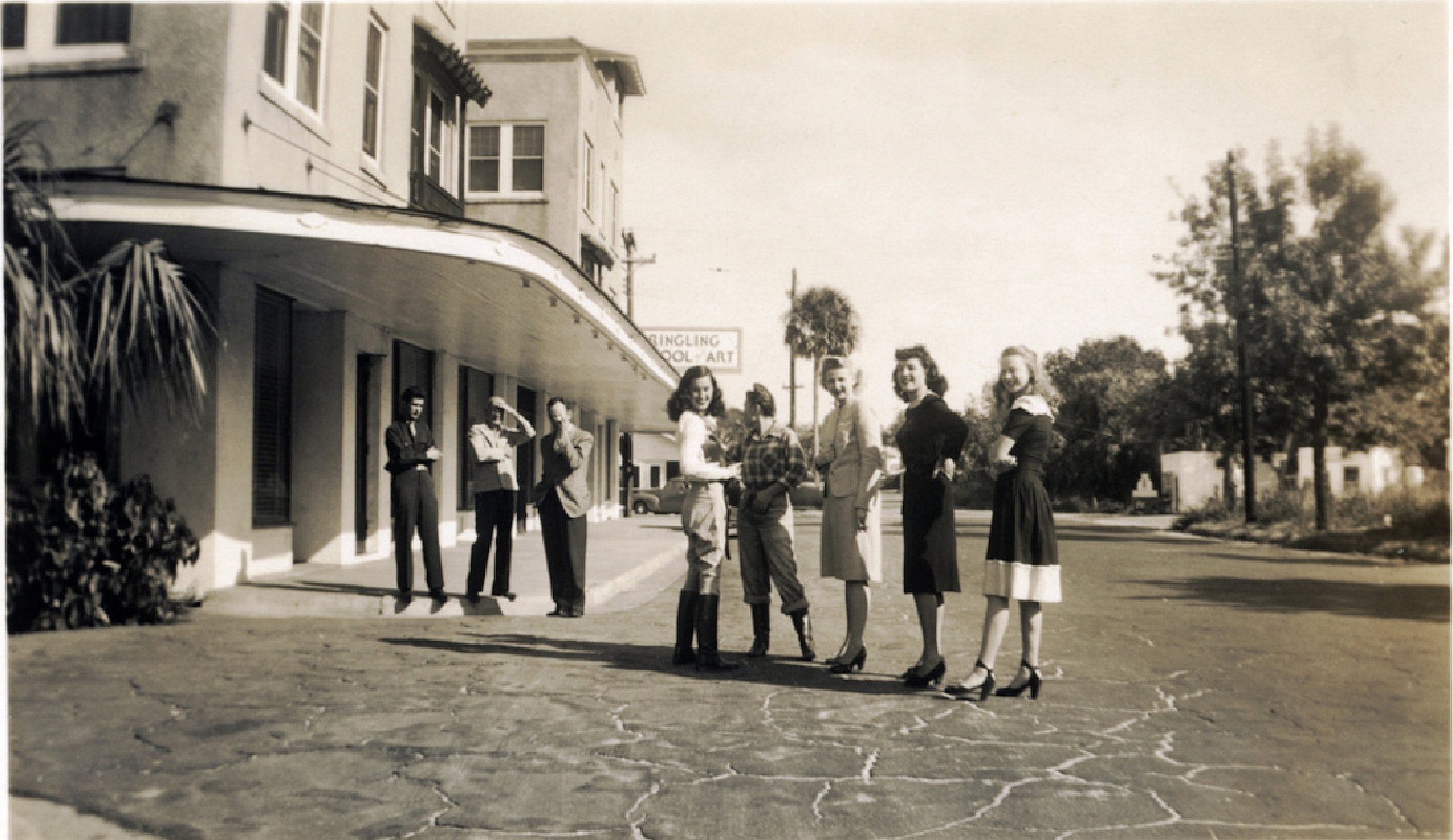 Black and white photo of seven women standing on a street corner, some wearing dresses with high heels, and three men standing near a building with a sign that reads 'Springling School of Art', in a vintage downtown setting.