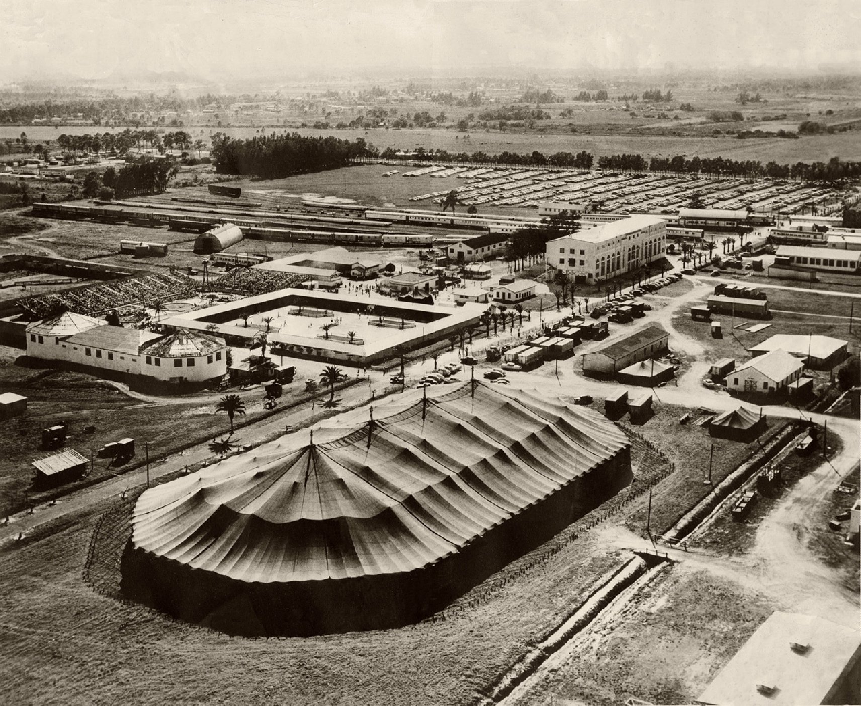 Aerial view of a large outdoor event with a tent, buildings, and surrounding area, black and white photograph.