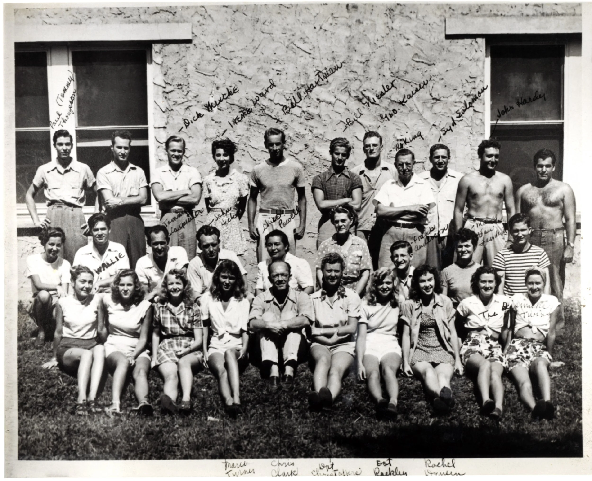 Black and white group photo of approximately 20 young adults and a few middle-aged individuals, some seated on the grass in front and others standing behind, in front of a stone building with two windows. The photo features handwritten names and notes overlayed, indicating identification of some individuals, with various casual clothing styles and some individuals shirtless, suggesting a summer or outdoor setting.