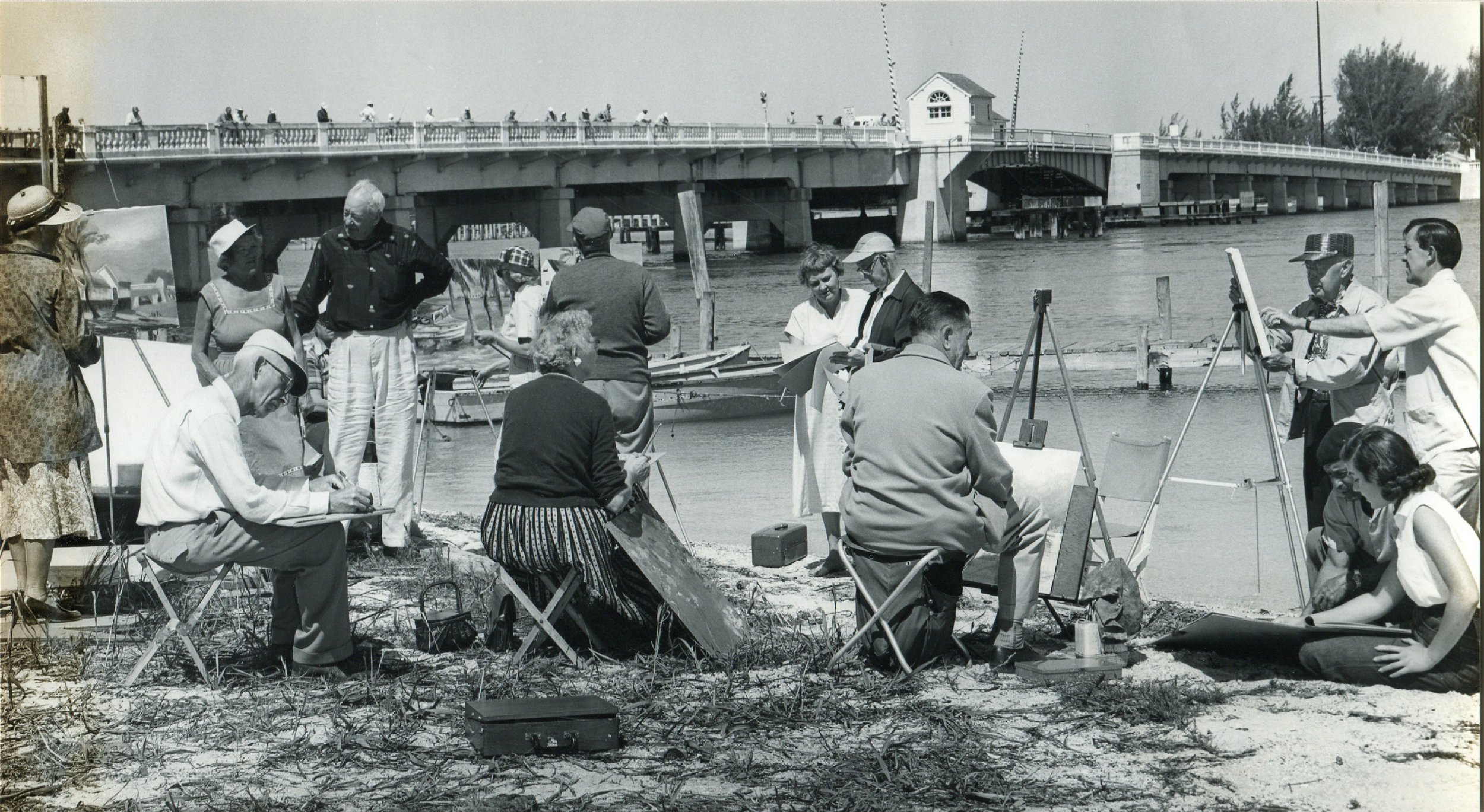 Group of people painting and sketching by a river under a bridge, with boats and a dock visible in the background.